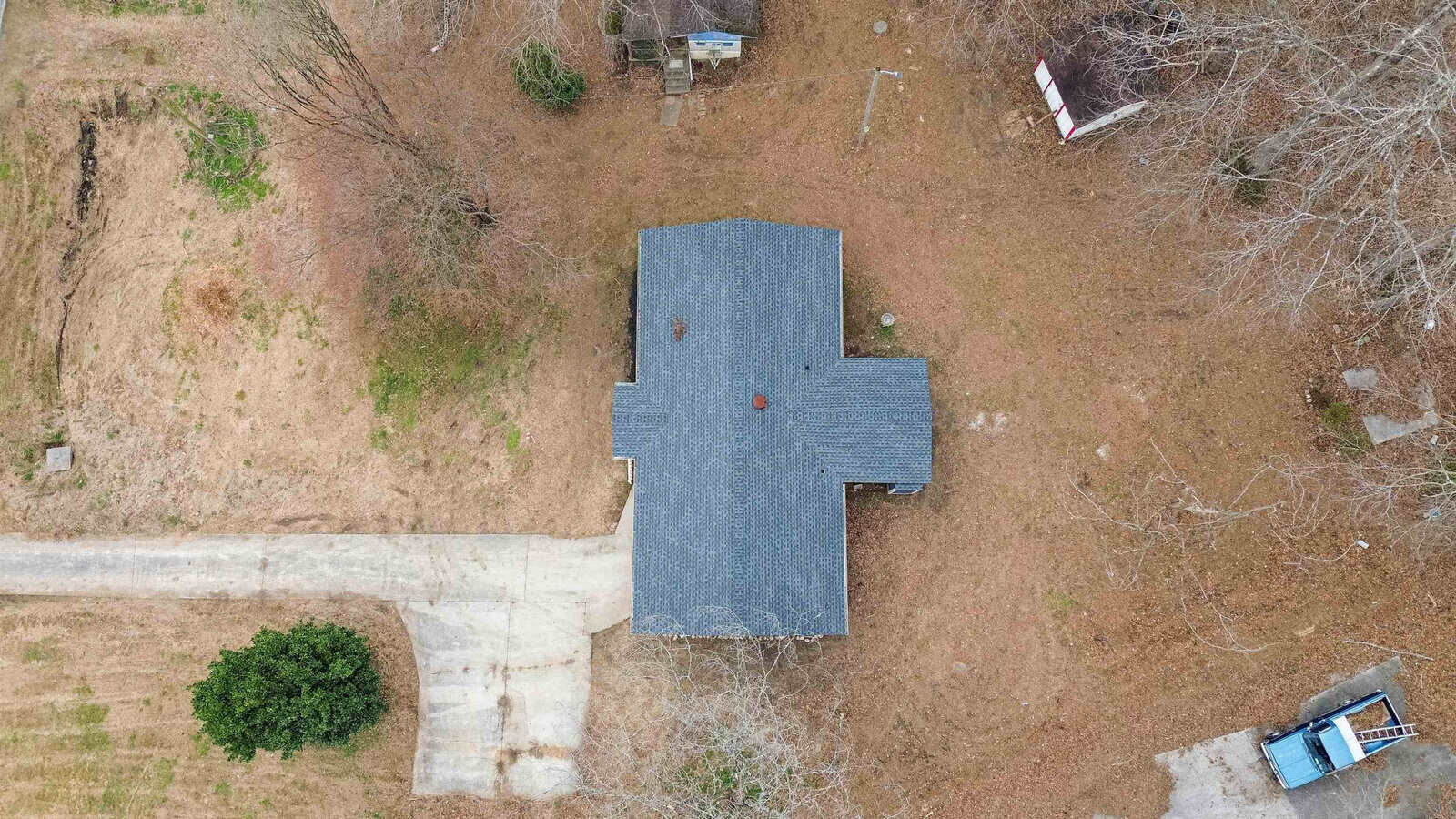 Aerial view of a small house with a dark gray roof surrounded by leafless trees and a brown lawn. A driveway leads to the house, and a blue truck is parked near the bottom right corner of the image.