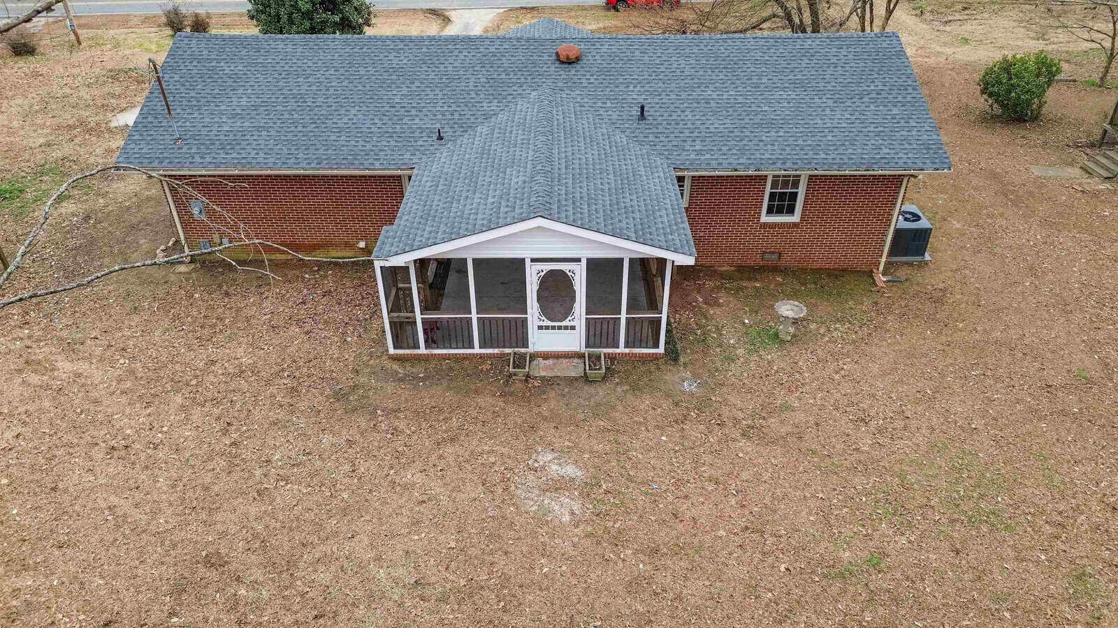 Aerial view of a single-story brick house with a dark gray shingled roof and a screened-in front porch. The yard surrounding the house is covered with light brown dried grass and sparse trees, indicating a winter or early spring season.