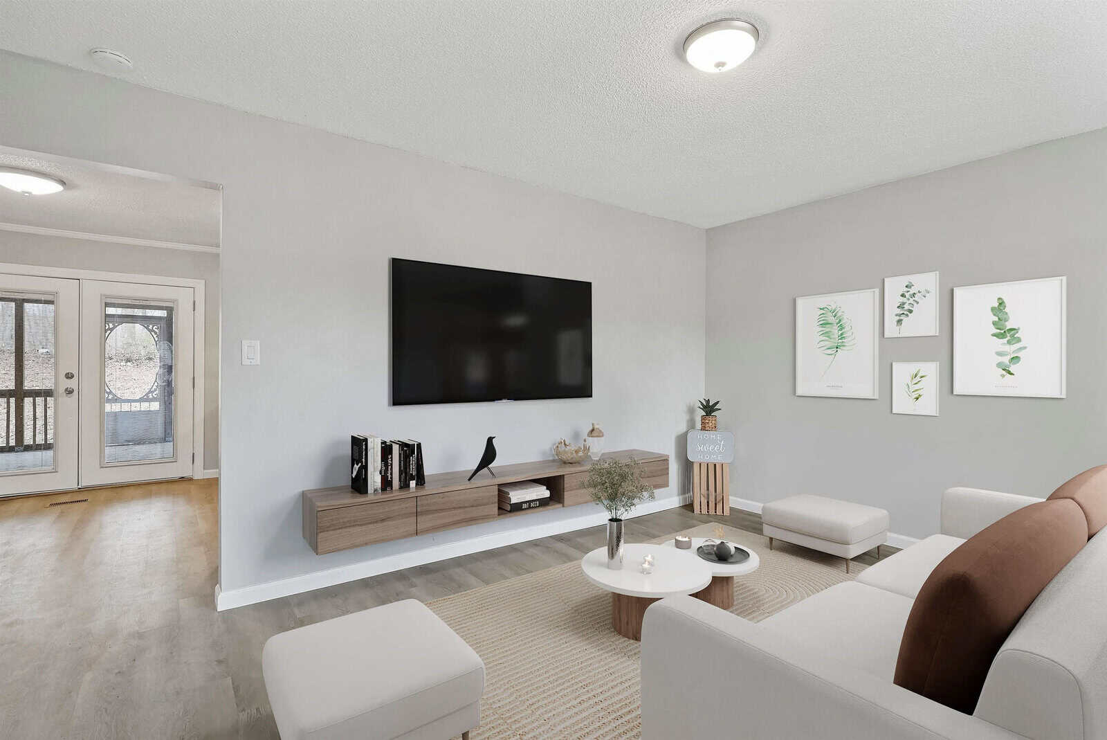 A modern living room with light gray walls features a wall-mounted flat-screen TV above a floating wooden console. The room includes a white and brown sofa set, a round white coffee table with plants, and framed botanical prints on the wall. The entryway is visible in the background.