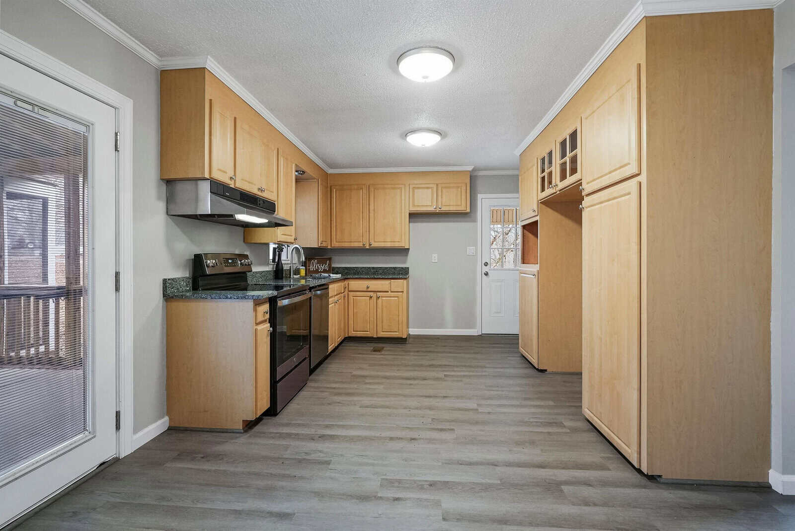 A modern kitchen with light wood cabinets, stainless steel appliances including a microwave and oven, and dark countertops. The room features gray flooring, white walls, a door with a glass panel on the left, and another door in the back.