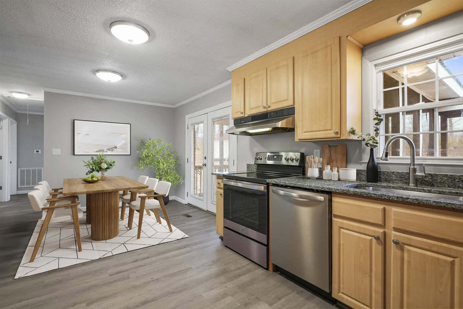 A modern kitchen with stainless steel appliances, light wood cabinets, and dark countertops. Adjacent is a dining area with a rectangular wooden table and white chairs on a geometric-pattern rug. Natural light enters through a window and sliding glass doors.