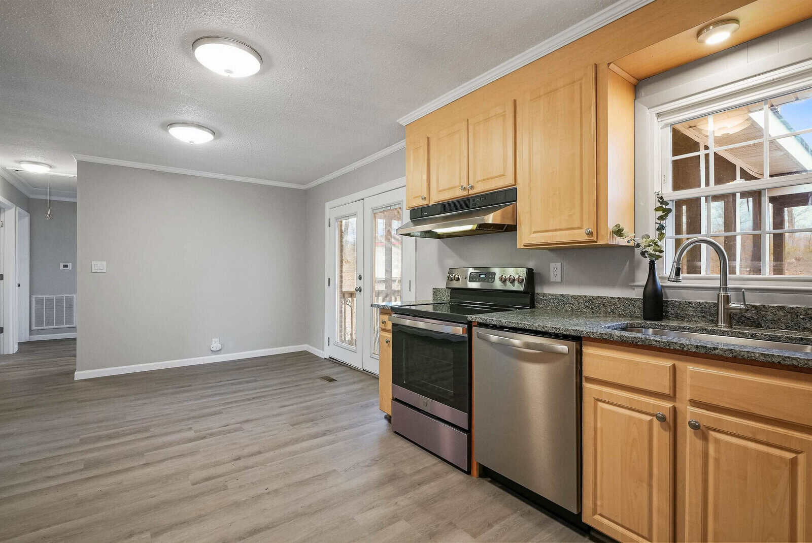 A modern kitchen with light wooden cabinets, stainless steel appliances including a stove and dishwasher, a granite countertop, and a window above the sink. The kitchen opens into an empty dining area with light grey walls and wood flooring. A white door leads outside.