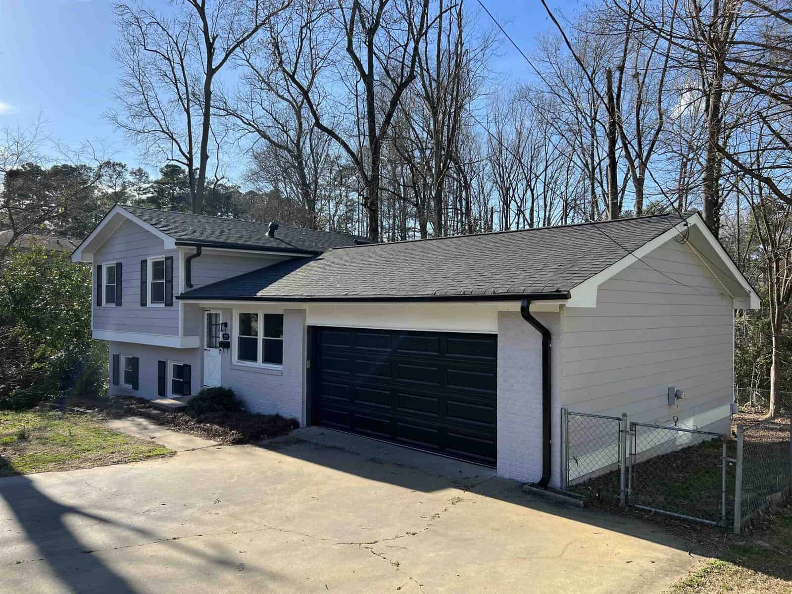 A split-level house with a black two-car garage door, featuring light blue sidings and white trim, situated on an incline with bare trees in the background under a clear sky.