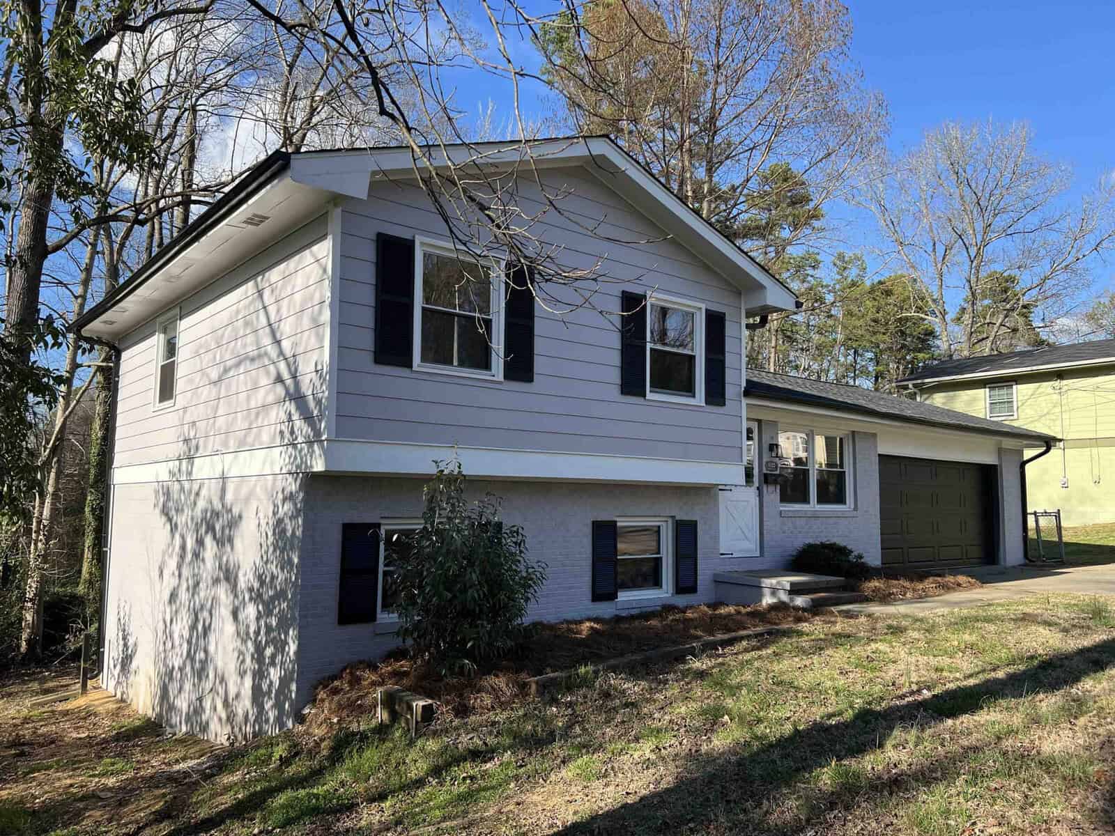 A two-story house with white siding, black shutters, and an attached single-car garage on a sunny day with trees in the background.