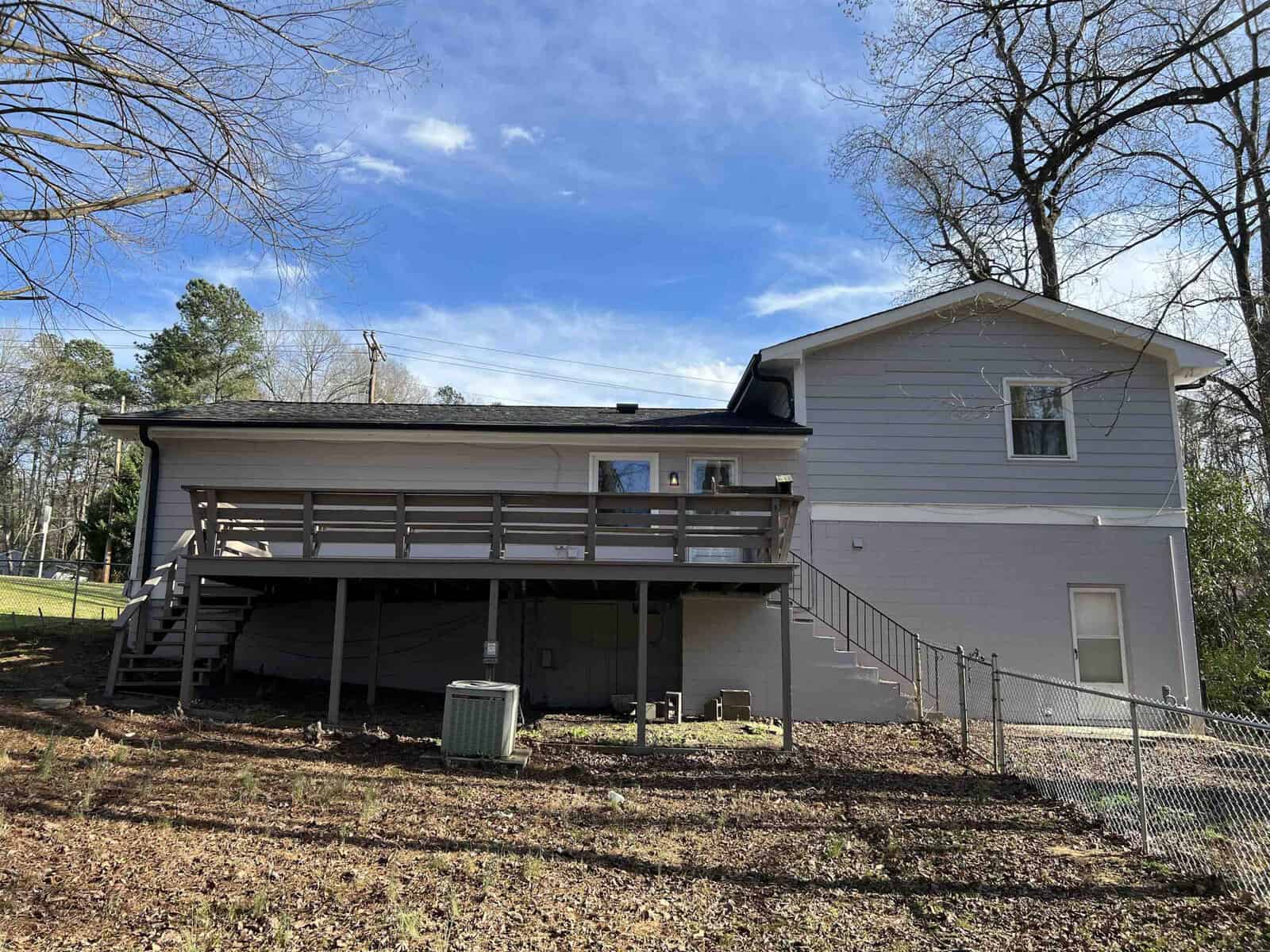 A two-story house with a gray exterior, featuring a large wooden deck and a chain link fence on a sunny day with trees in the background.