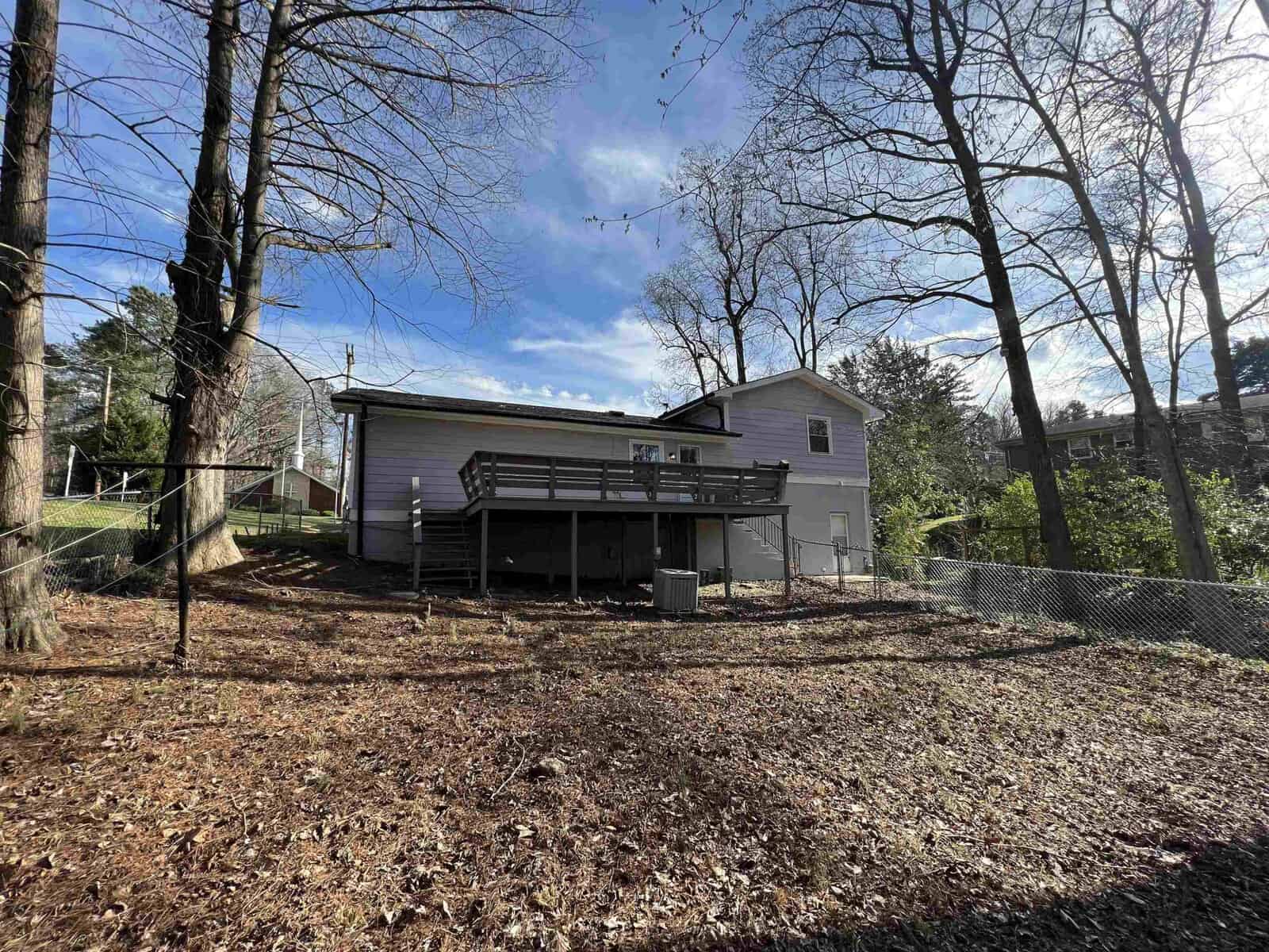 A backyard view of a two-story house with a raised deck, surrounded by leafless trees under a partly cloudy sky.
