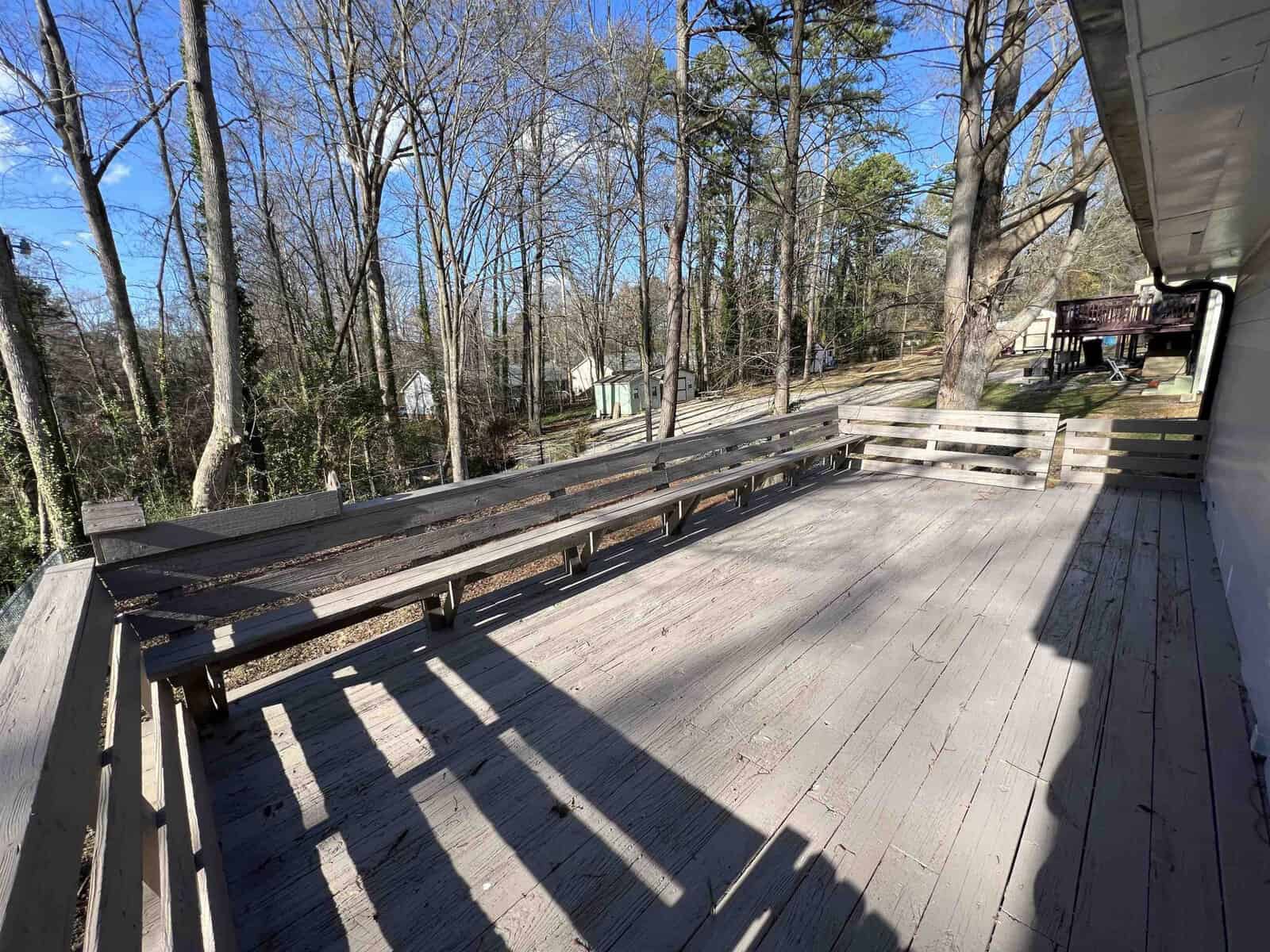 A spacious wooden deck with built-in benches bathed in sunlight, surrounded by leafless trees with a clear blue sky overhead.
