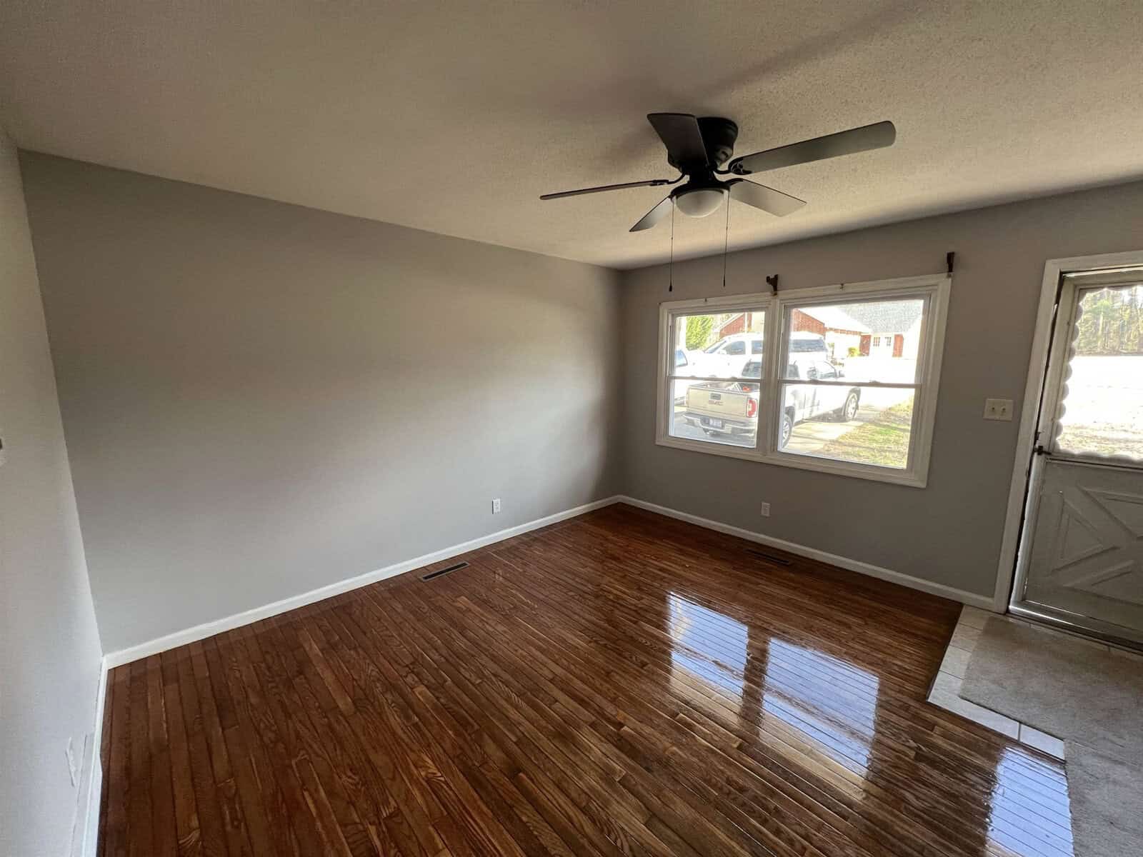 An empty room with glossy hardwood floors, a ceiling fan, and a window providing a view of the outside neighborhood.