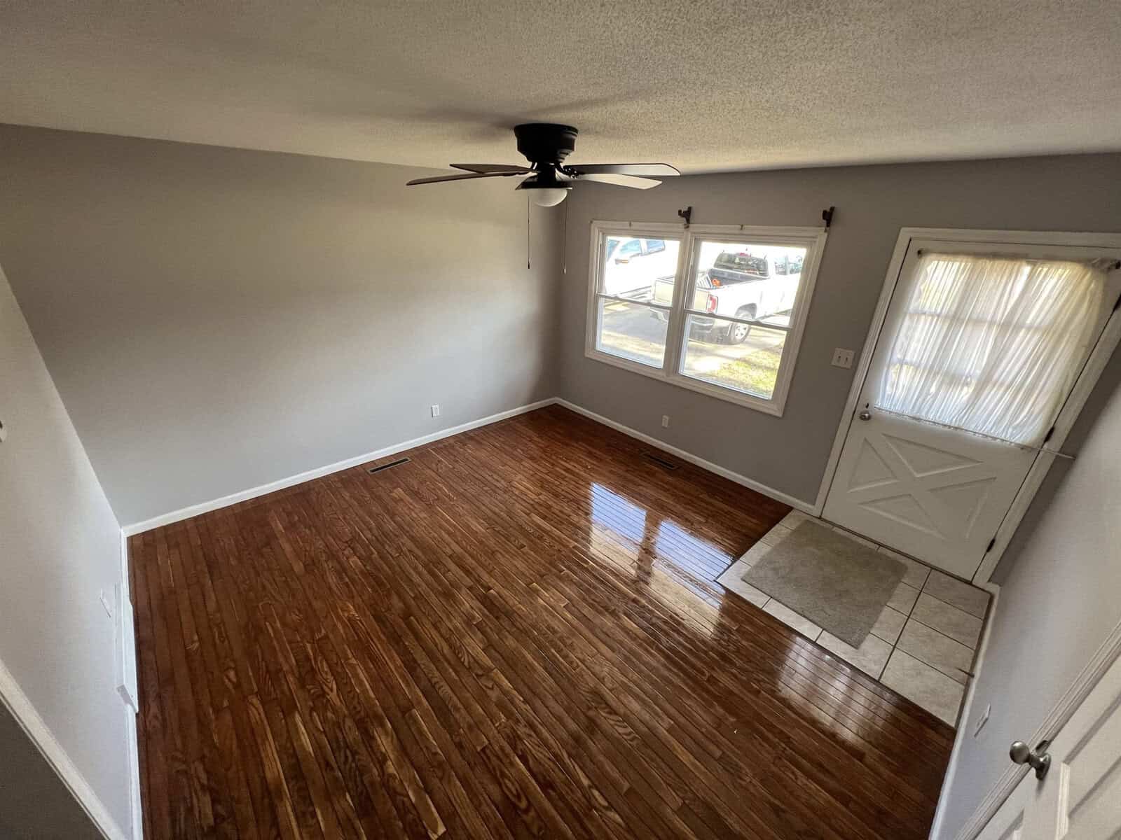 An empty room with polished wooden flooring, a ceiling fan, a closed white door to the left, and a window with blinds partially down on the right wall.