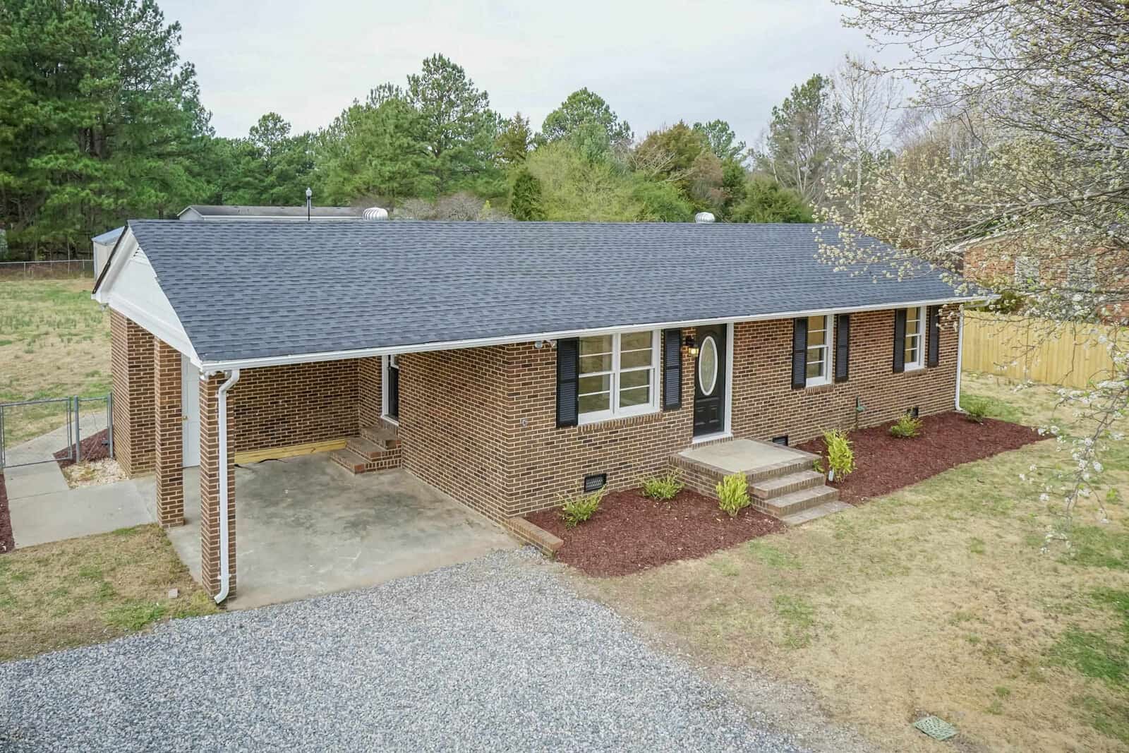 A single-story brick house at 1064 Abbott Road, Henderson NC, with a gray roof and a gravel driveway, surrounded by trees and a lawn.