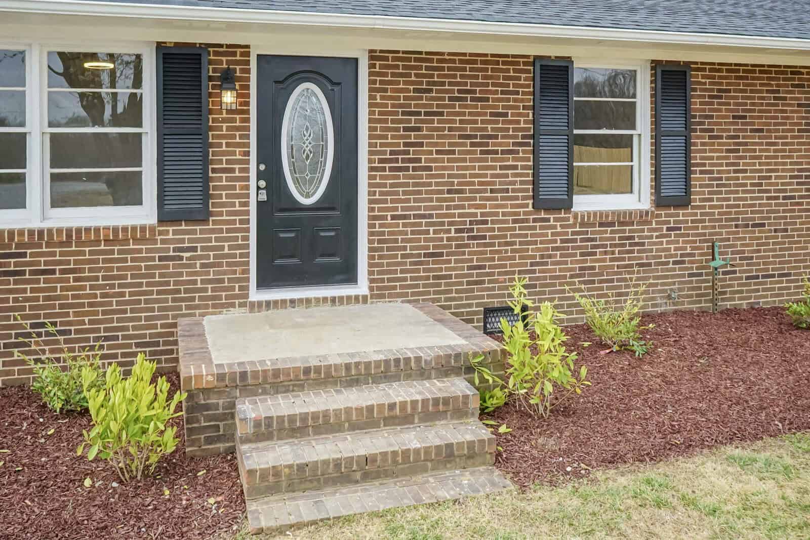 A brick house with a black front door flanked by windows with black shutters, featuring a small porch with concrete steps, and a landscaped area with young plants and mulch.