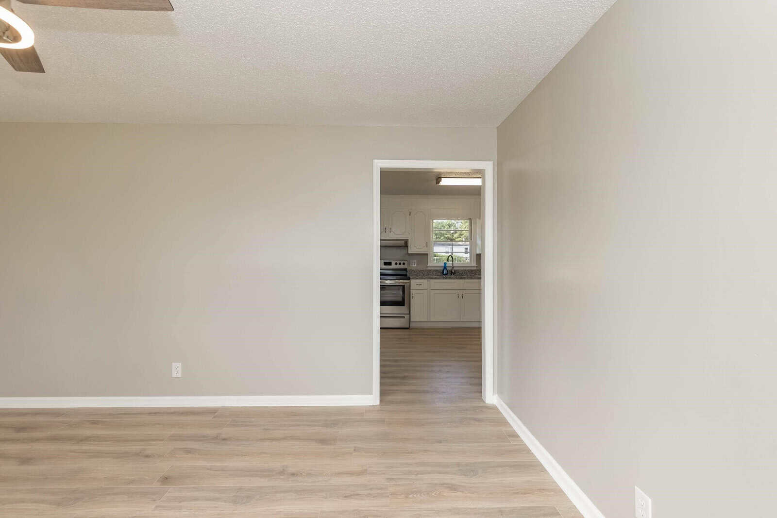 Empty room with laminate flooring leading to a kitchen area with white appliances.