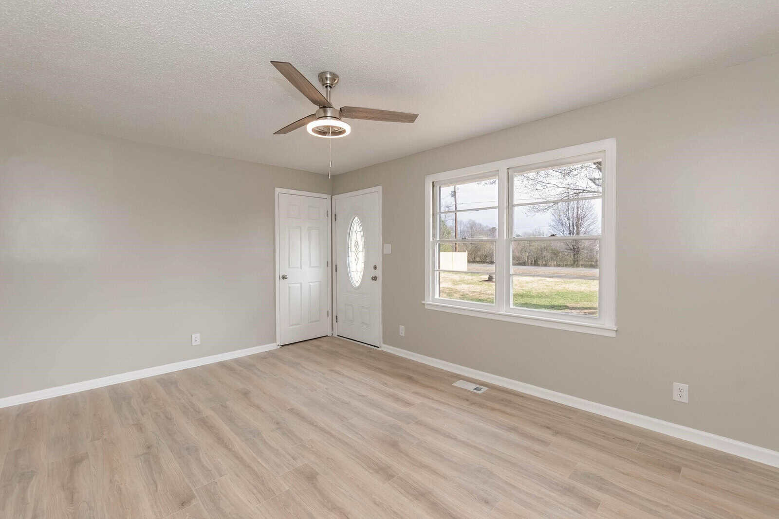 An empty room with beige walls, wooden flooring, a ceiling fan, and two windows providing a view of the outdoors.