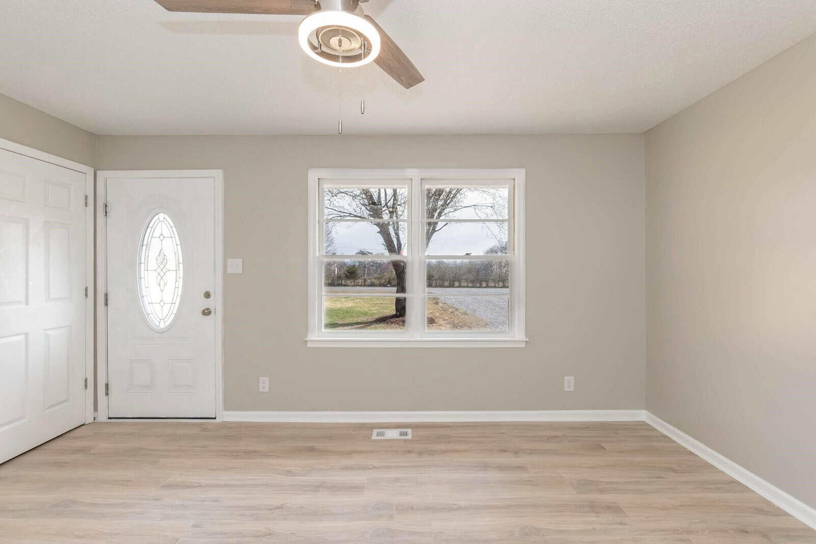 An empty room with beige walls and laminate flooring, featuring a front door with ornamental glass and a large window with a view of the outdoors.