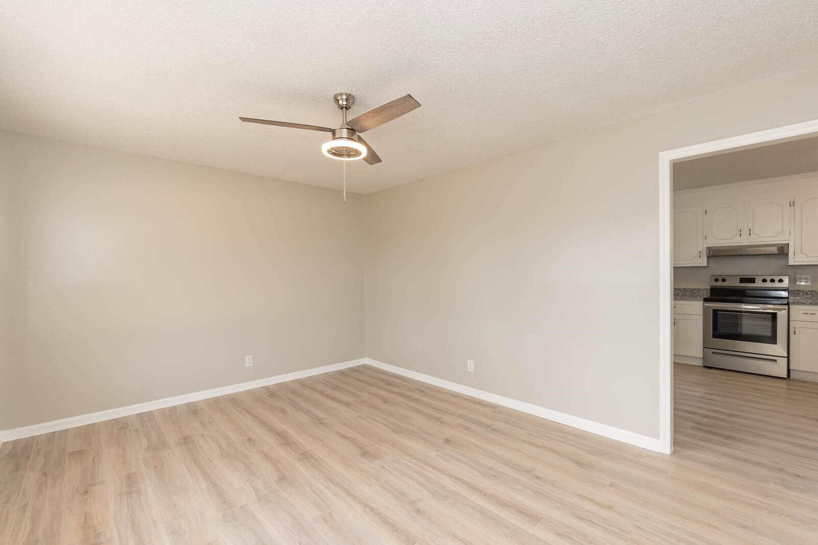 An empty room with beige walls and laminate flooring features a ceiling fan and an open doorway leading to a kitchen with white appliances.