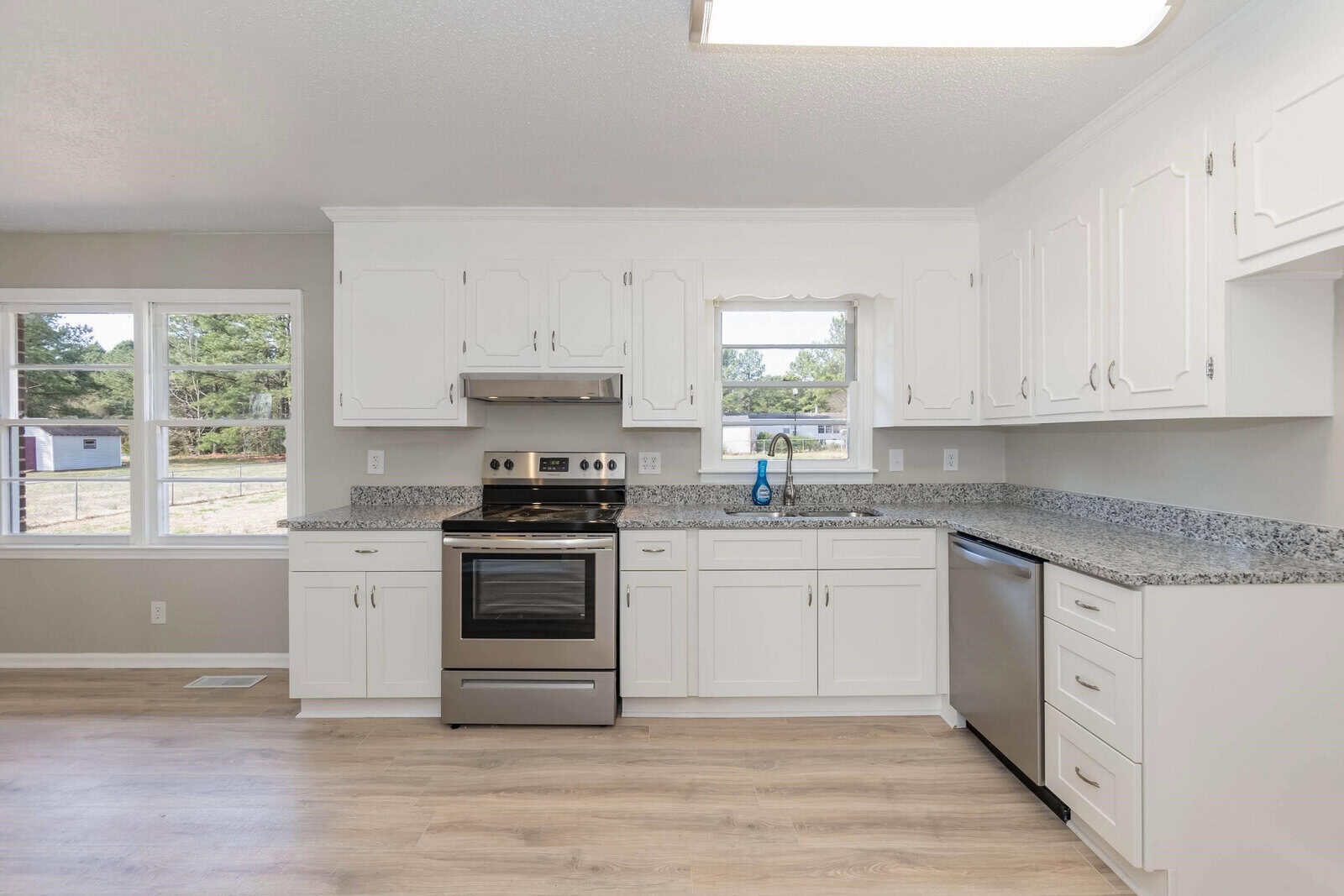 A modern kitchen interior with white cabinetry, granite countertops, and stainless steel appliances, featuring a window above the sink and vinyl plank flooring.