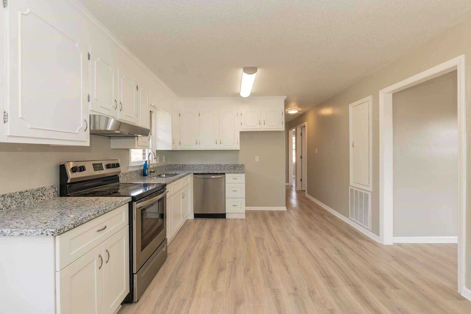 Spacious kitchen featuring white cabinetry, stainless steel appliances, granite countertops, and wood-style flooring, with an adjacent hallway.