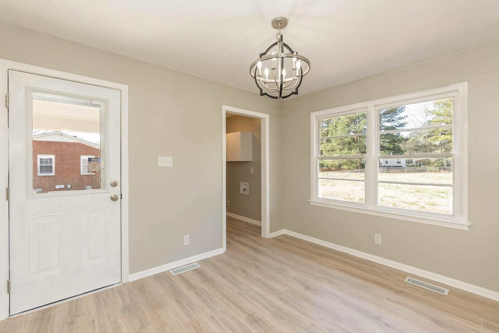 An empty room featuring a chandelier, with laminate flooring, a white entrance door on the left, and a large window with a view of the outdoor area.