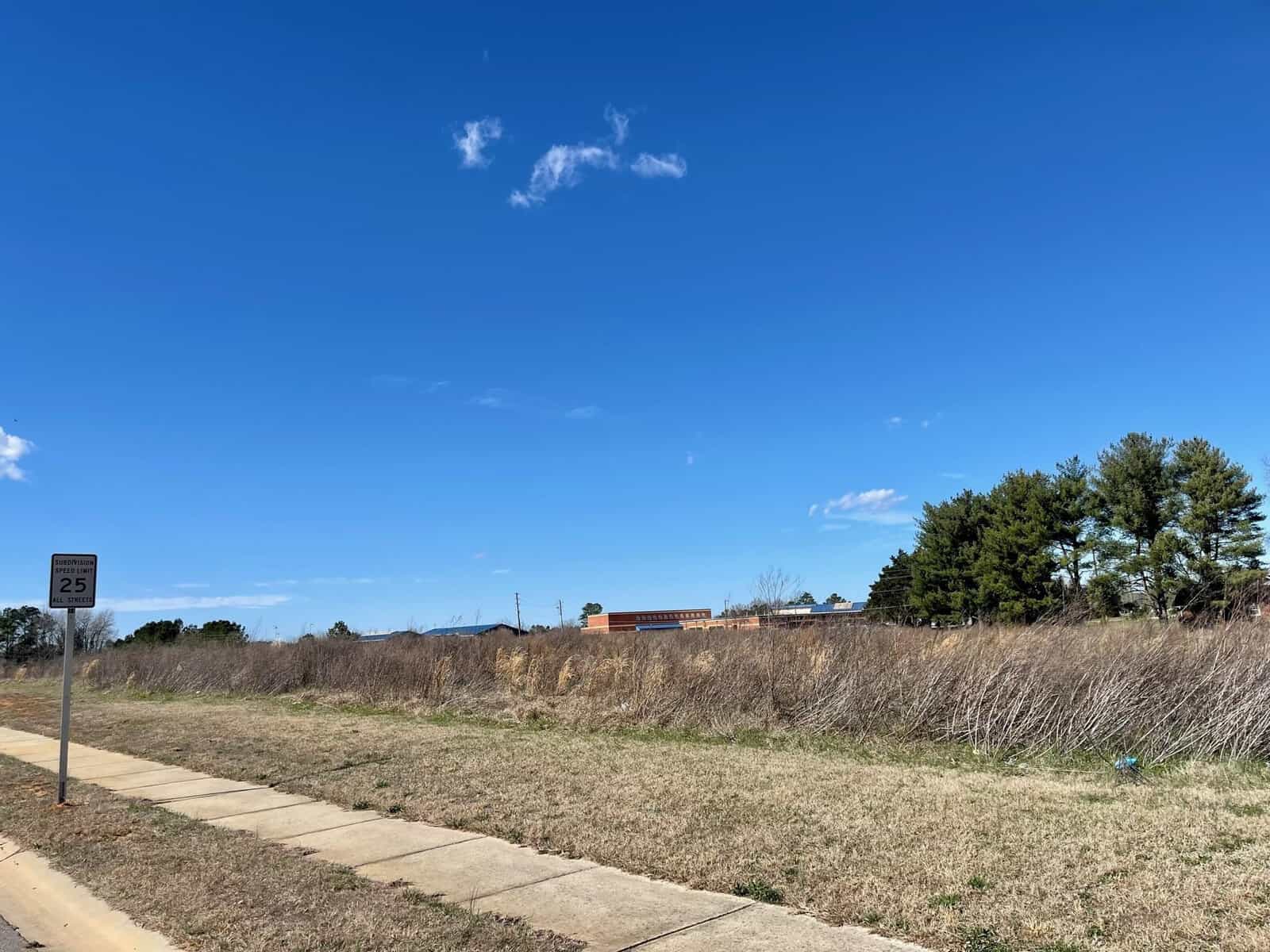 A suburban landscape at 000 Ashmont Lane, Henderson NC, featuring a paved sidewalk alongside a grassy, undeveloped plot of land under a clear blue sky, with scattered trees and a distant