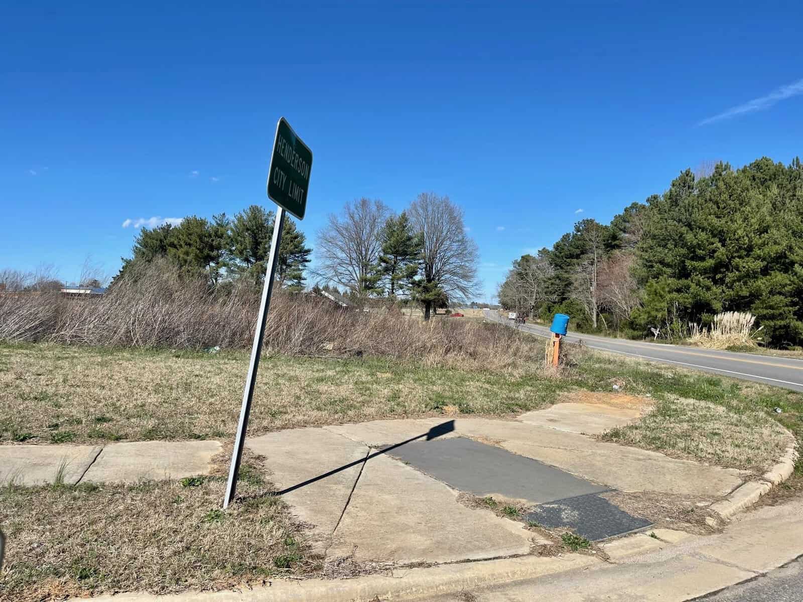 A road sign titled "nc train" by a stopgap walkway on a grassy patch with trails, alongside a road under a clear blue sky. trees and a distant traffic light are visible in the background.
