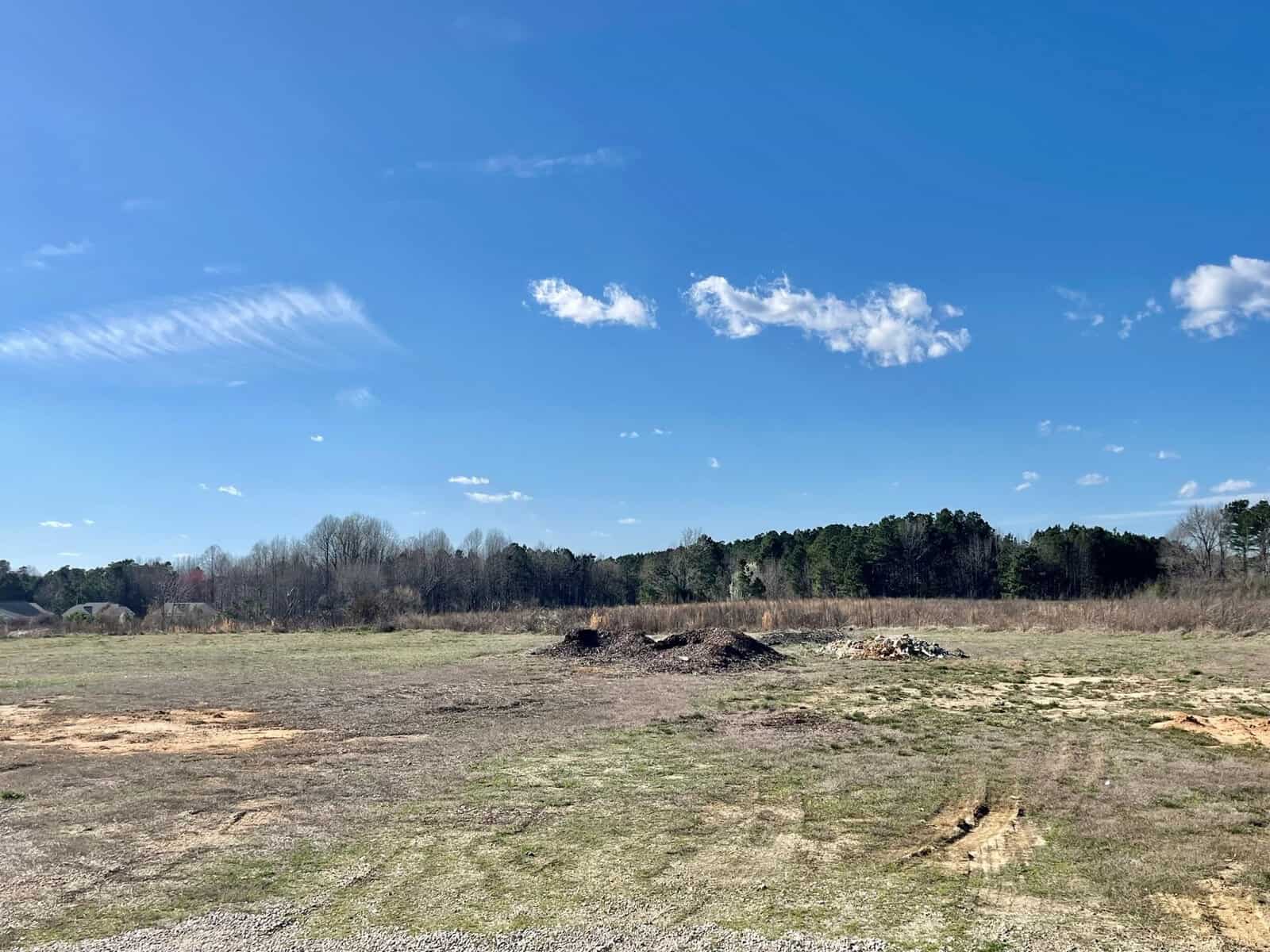 A wide open field with sparse grass and patches of dirt under a clear blue sky with a few scattered clouds, surrounded by distant trees and some debris on the ground.