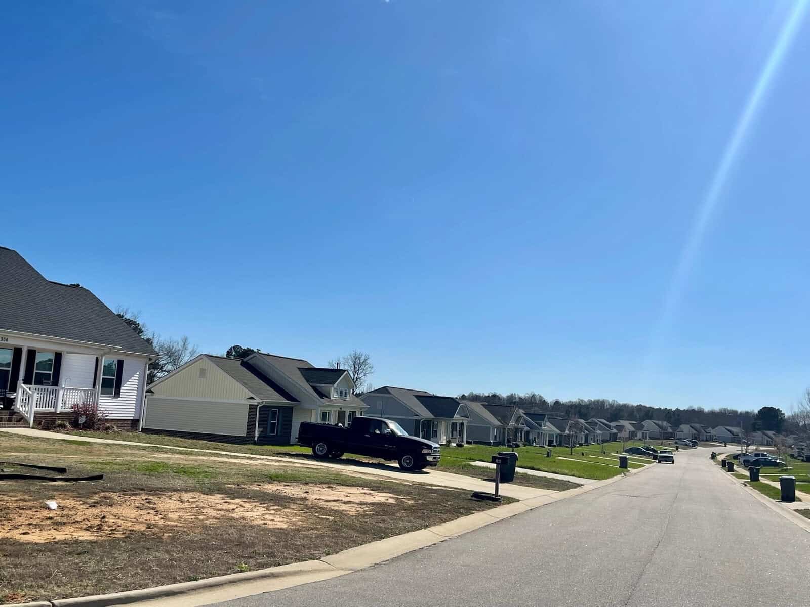 A sunny suburban street with single-family homes and a black pickup truck parked in a driveway. the sky is clear blue with a bright sunbeam on the left.