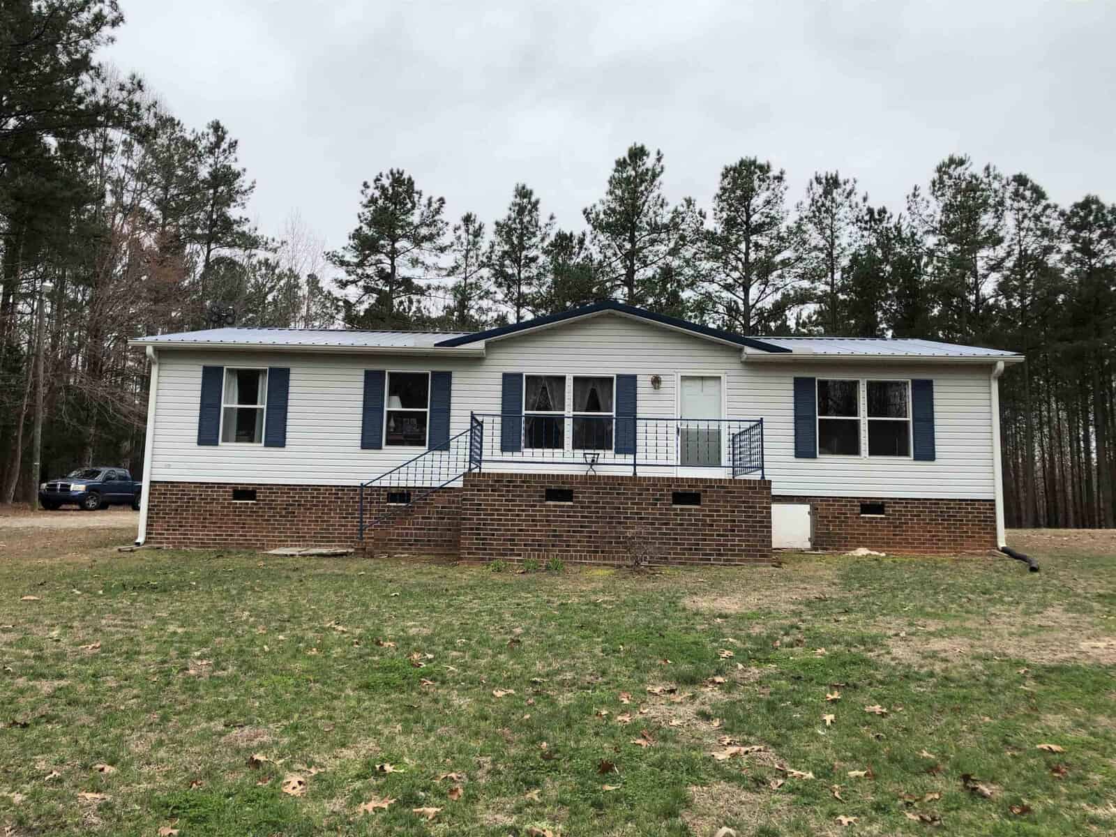 A white single-story house at 129 Jamel Lane, Henderson NC, with a raised foundation, featuring a central entrance with steps and a railing, situated on a lawn with scattered trees in the background