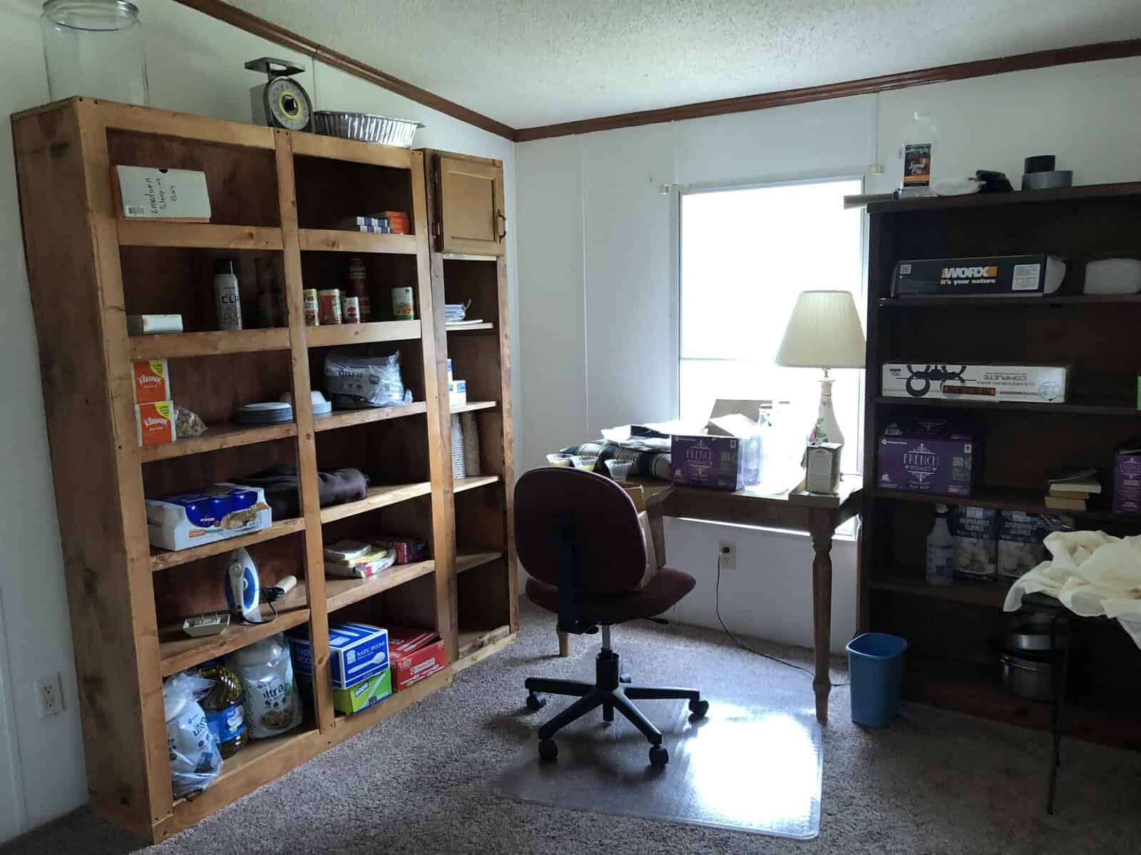 A home office with wooden shelves stocked with various items, an empty chair in front of a desk, and miscellaneous objects scattered throughout the room.