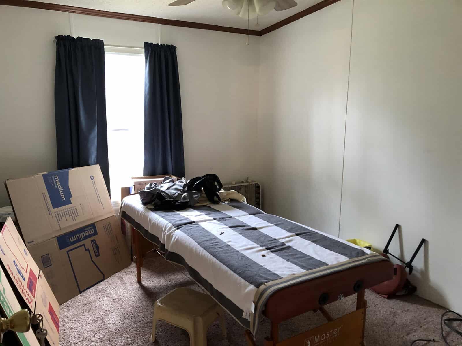 A bedroom with a single bed covered in a striped blanket, a pair of dark curtains on the window, some clothes on the bed, a cardboard box on the left, and a vacuum cleaner on the right.