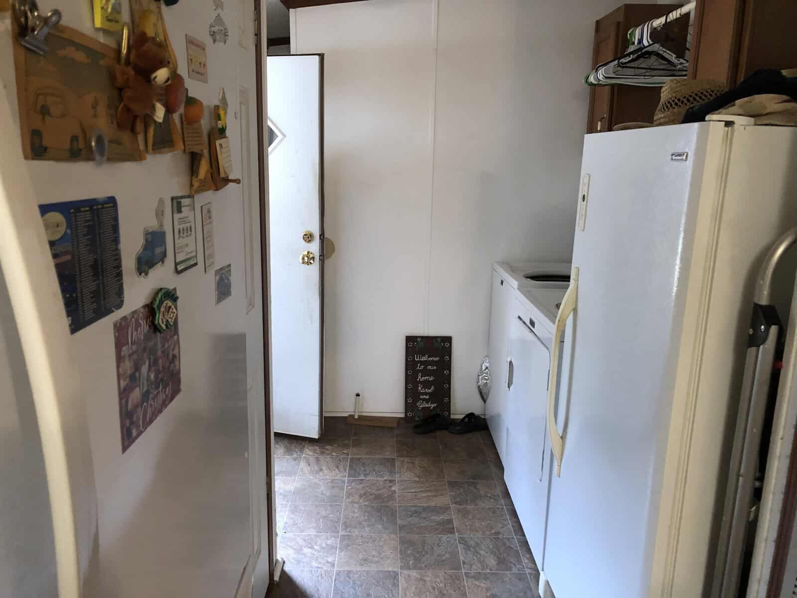 A residential kitchen with a refrigerator on the left, adorned with magnets, a dryer in the center, and a pantry door to the right. the floor is tiled, and there are various objects and appliances on the countertops.