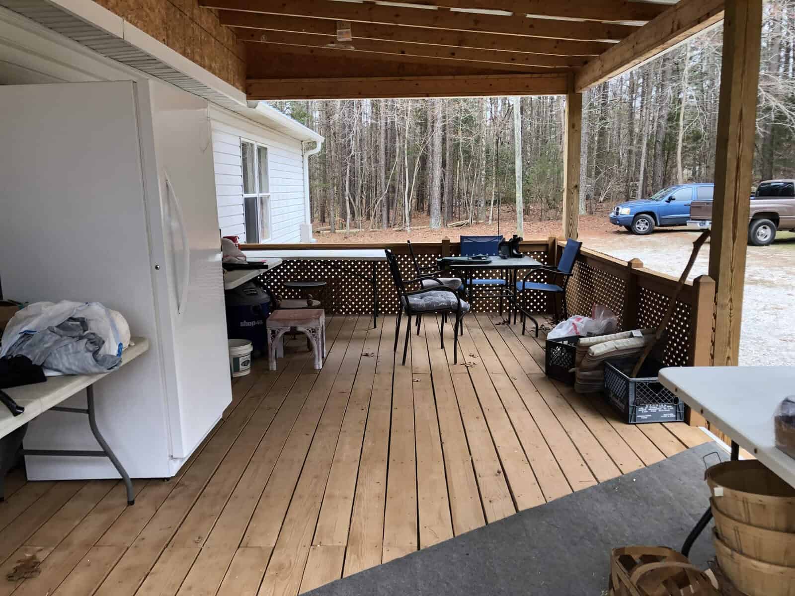 A spacious wooden porch with household items and furniture, overlooking a yard with a blue pickup truck parked in the background.