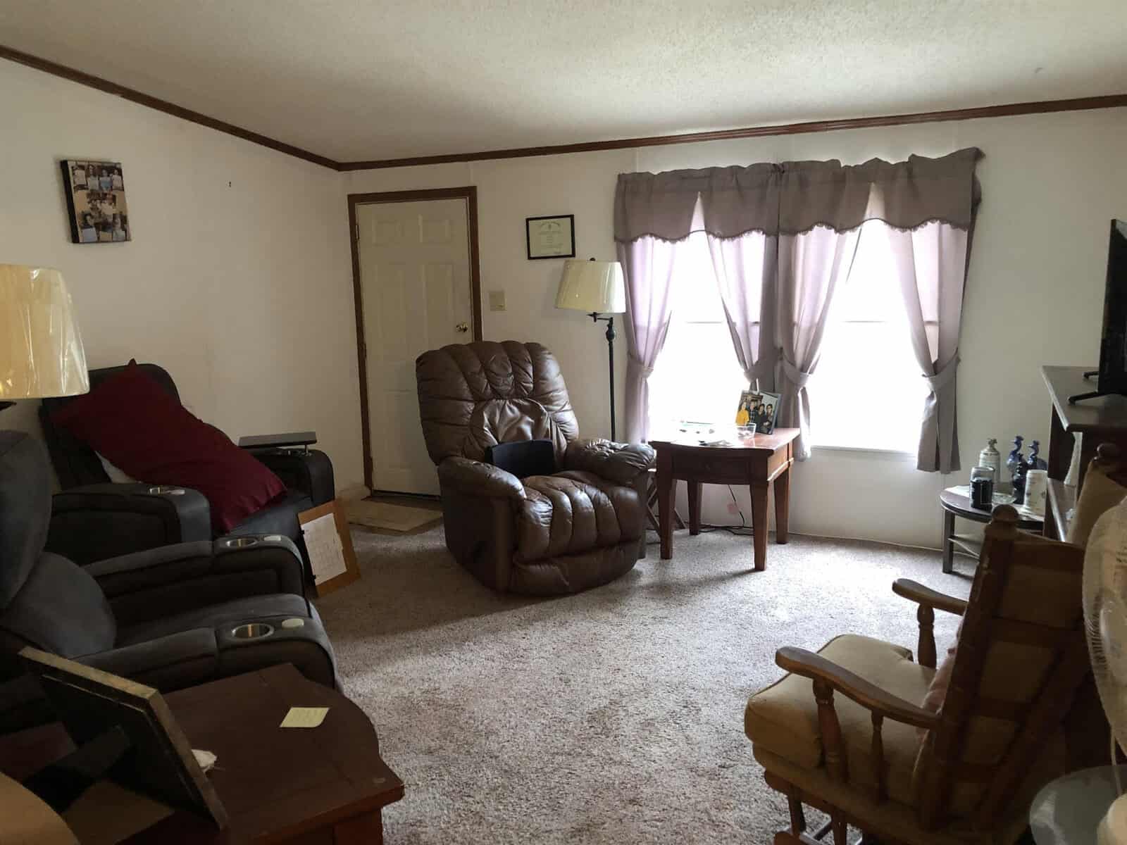 A tidy living room with a recliner, sofas, a side table with a lamp, and a window dressed with sheer curtains.