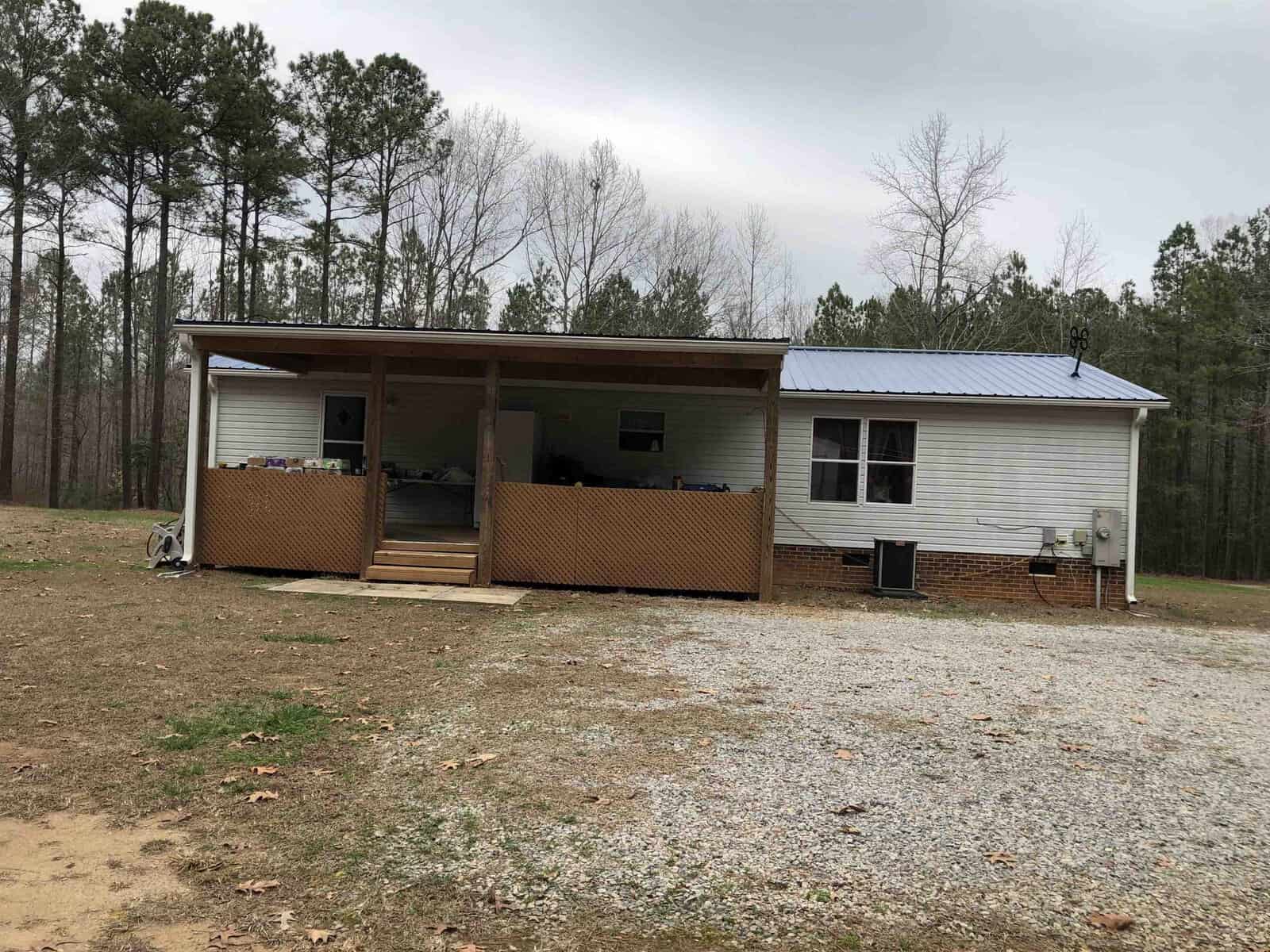 A single-story house with a covered porch and partially wood latticed base, featuring an hvac unit on the side, under a cloudy sky. the ground in front is covered with gravel, and there are trees in the background.