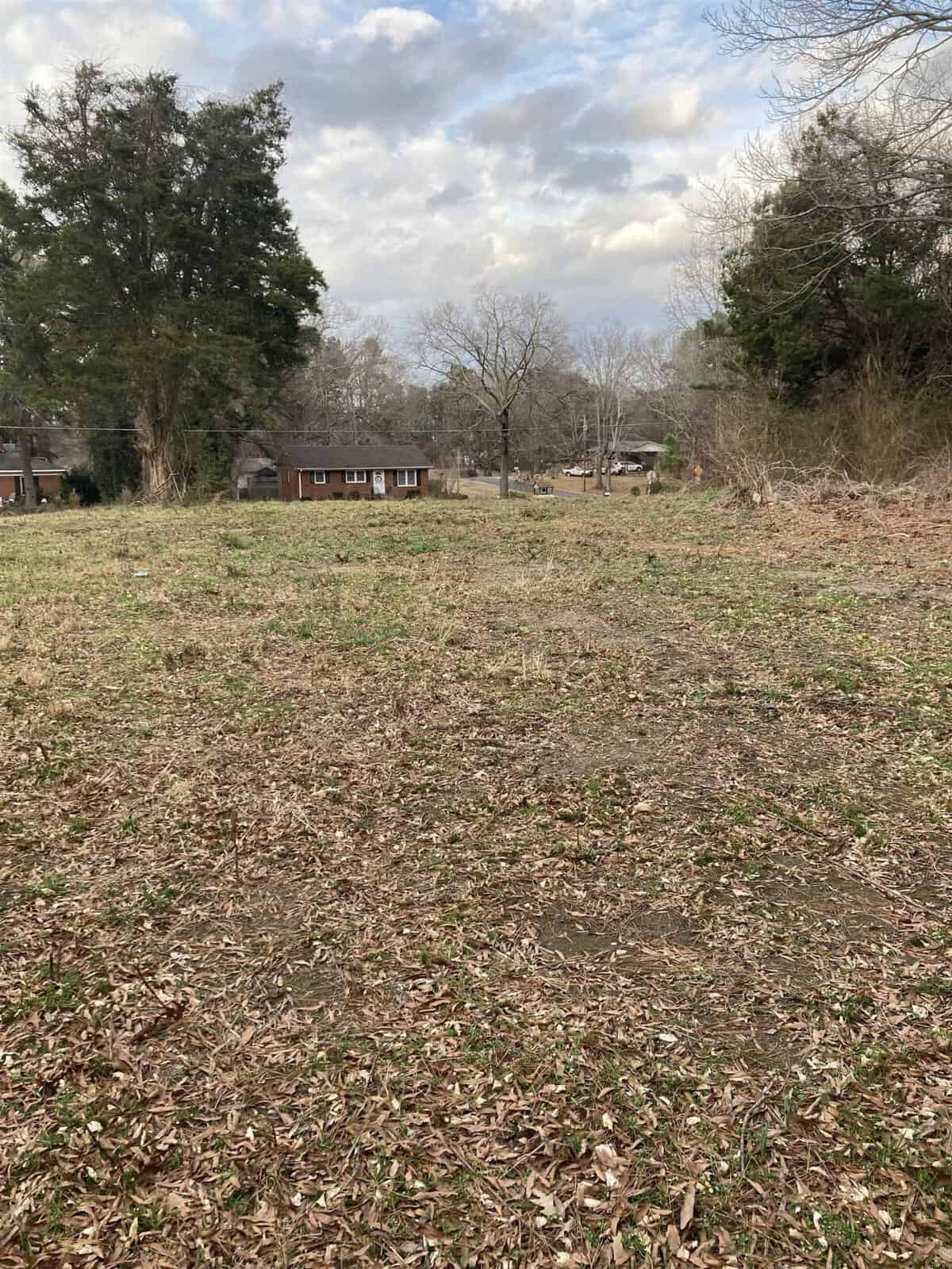 A vacant lot with scattered leaves and sparse vegetation, with a residential house and trees in the background under an overcast sky.