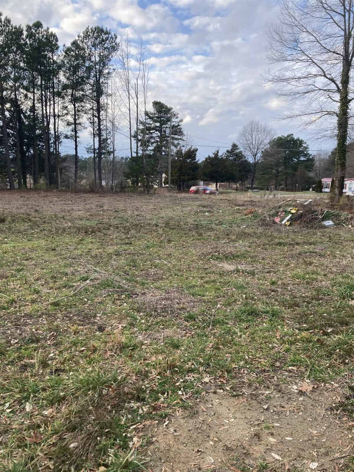 A sparsely vegetated field with scattered debris and a backdrop of trees under a cloudy sky.