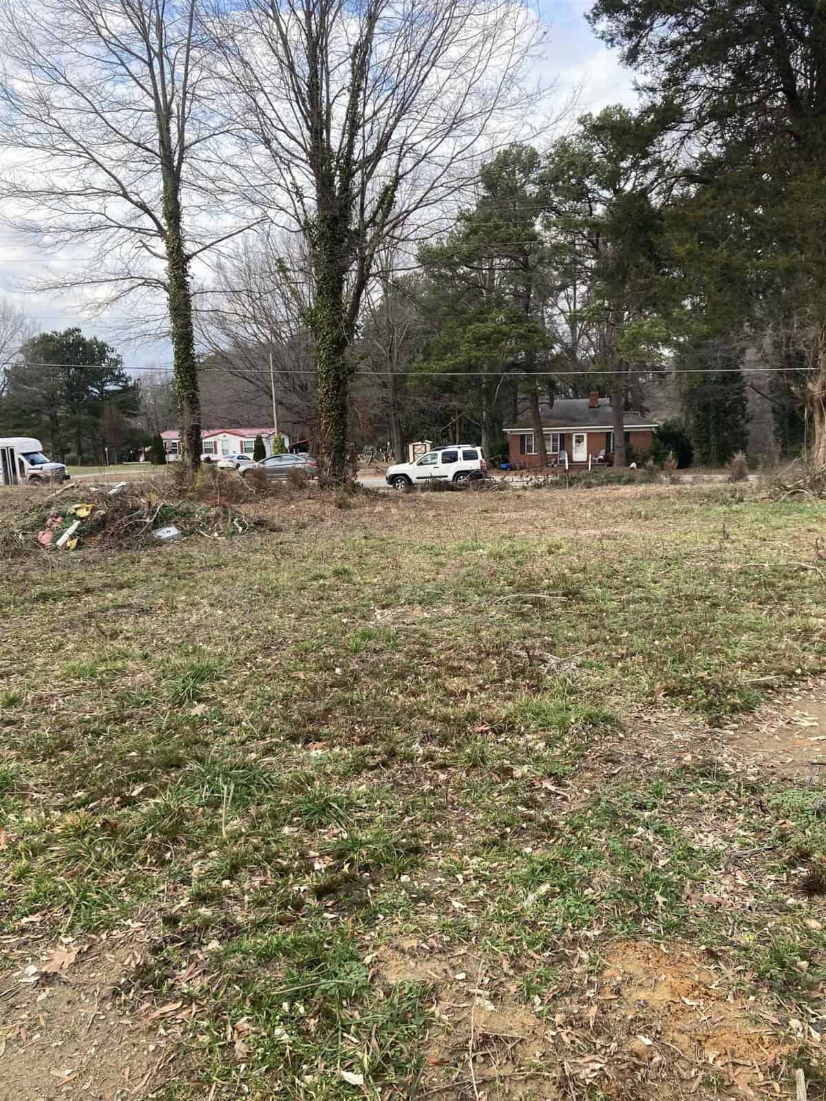 An open grassy area with trees and scattered debris, with a residential neighborhood and parked vehicles in the background.