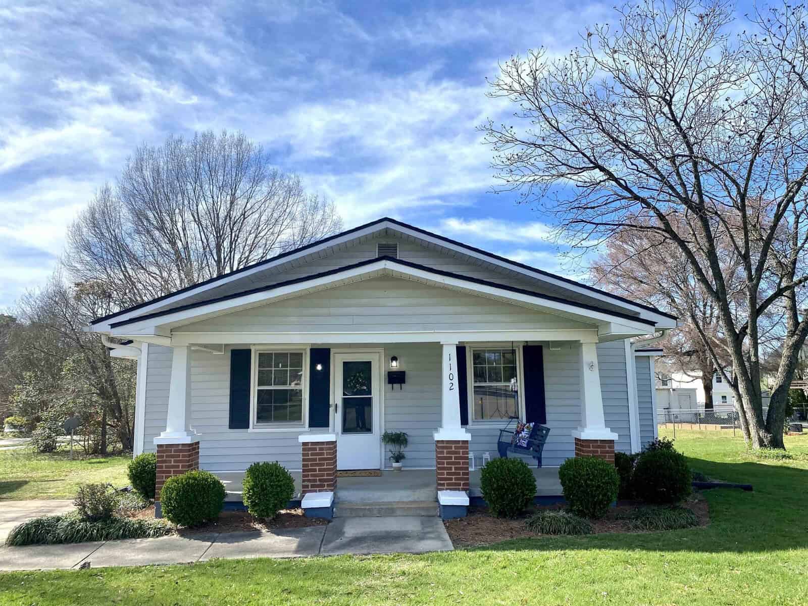 A small, single-story house with white siding and a front porch on a sunny day, with a neatly manicured lawn and bare trees in the background.