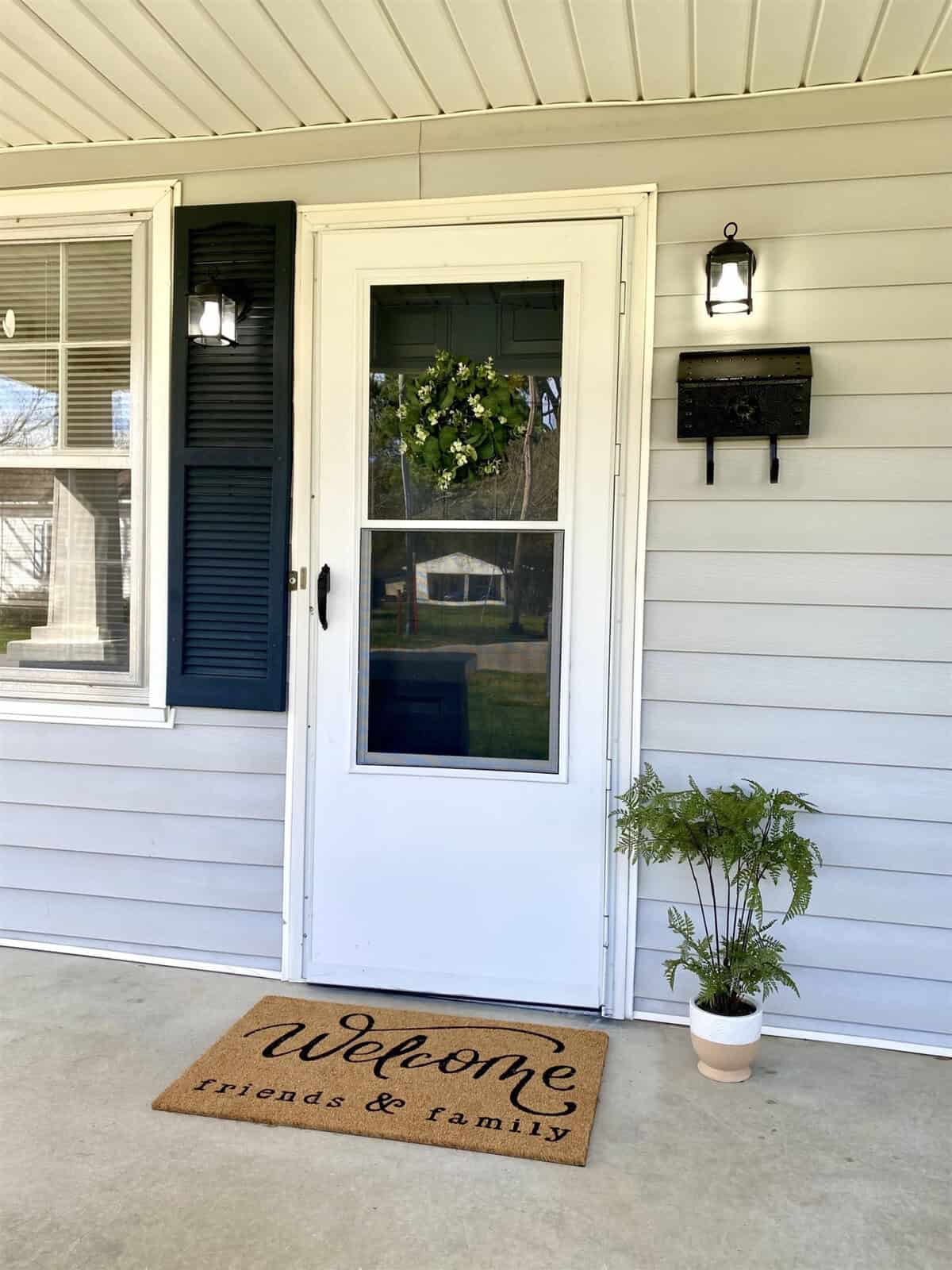 A welcoming white front door with a glass insert, adorned with a green wreath and flanked by a potted plant, with a doormat reading "welcome friends & family" placed on the porch.