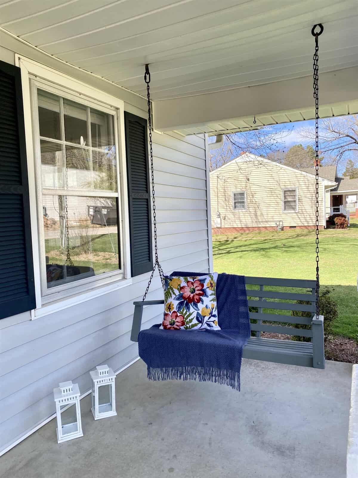 A porch with a swing bench adorned with a blue throw blanket and a patterned cushion, complimented by two white lanterns on the floor.