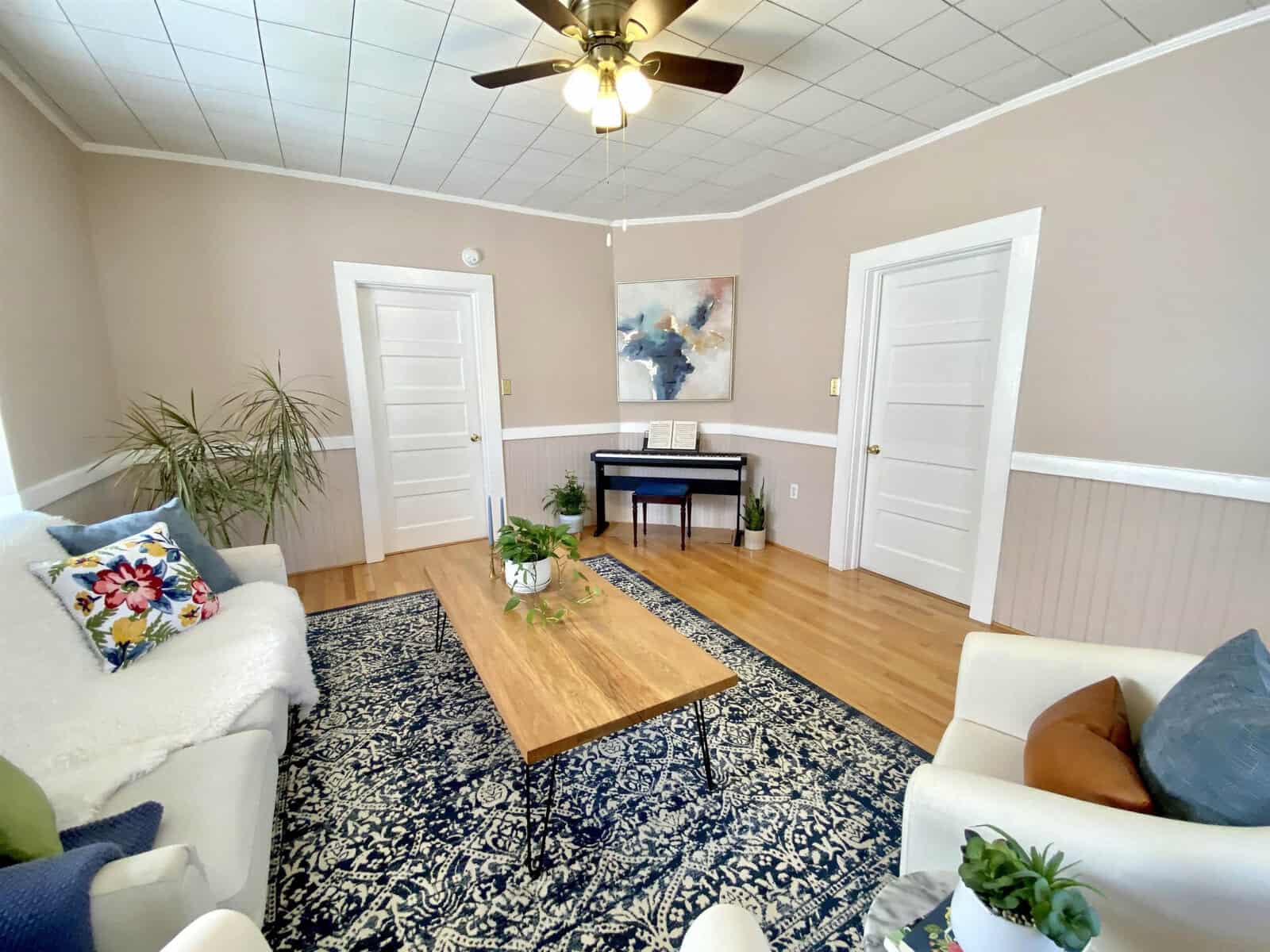 A spacious living room with hardwood floors, furnished with a beige sofa, two armchairs, a wooden coffee table, and a decorative rug. the room features a piano against a wall, two white doors, and a ceiling fan, with a large abstract painting as a focal point.