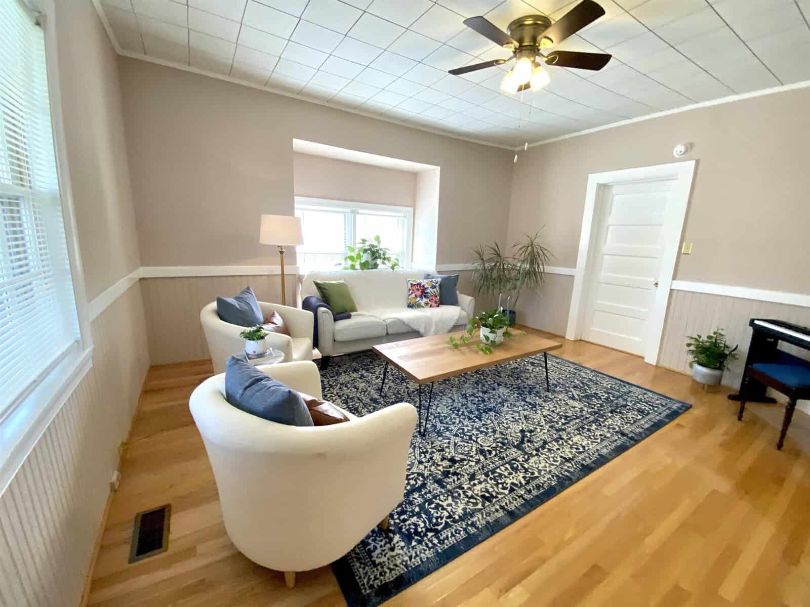 A spacious living room with neutral walls and hardwood floors, featuring a couch, two armchairs, a coffee table, and a ceiling fan. a plant adds a touch of greenery to the corner of the room.