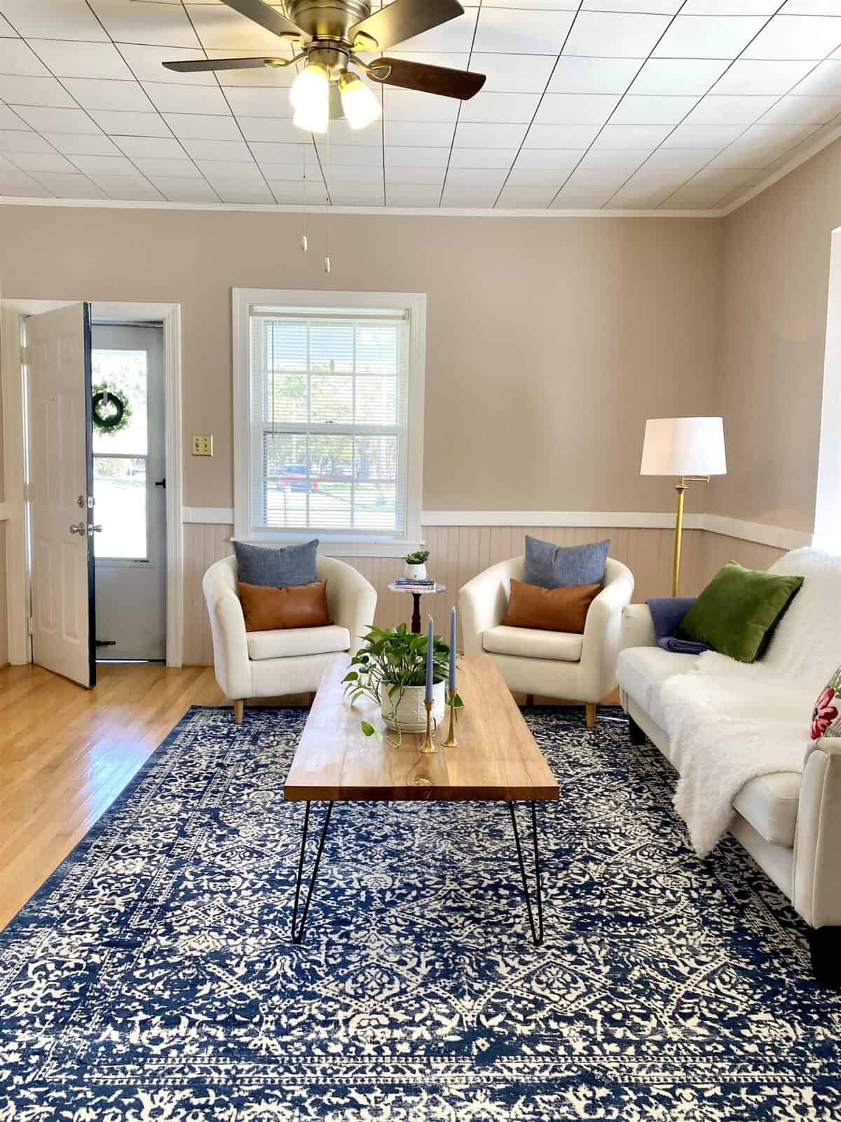 A well-lit living room with two white armchairs, a wooden coffee table, and a large blue area rug. a ceiling fan is visible above, and a lamp stands beside a window with white trim. the room has hardwood floors and a white front door with a decorative wreath.
