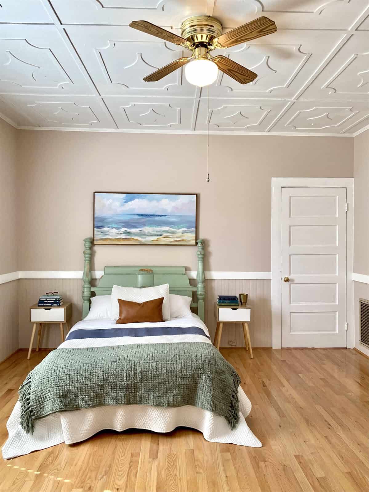 A neatly arranged bedroom with a mint green bed frame, green and white bedding, side tables, a ceiling fan, and a landscape painting above the bed.