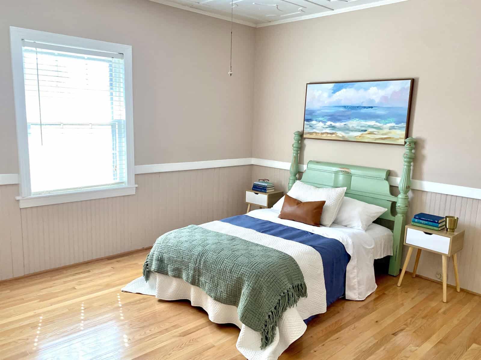 A well-lit bedroom with a seafoam green twin bed, a blue and white bedspread with a green throw, a framed ocean painting above the bed, wood flooring, and a small side table by the window.