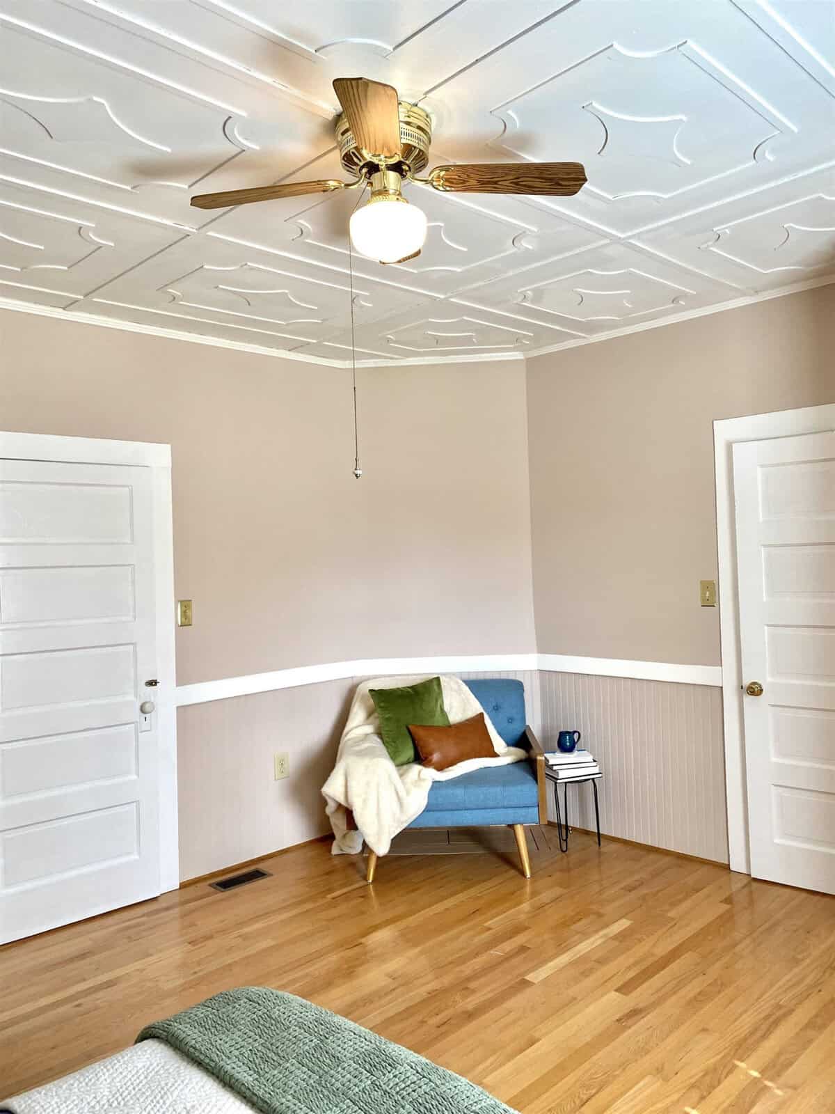 A cozy corner in a room with cream-colored walls featuring a stylish blue love seat adorned with a white throw and a single green pillow. the room boasts an elegant white ceiling with decorative patterns, complemented by a wooden floor and a classic ceiling fan with light fixture above. two white doors flank the seating area, one closed and the other slightly ajar, while a small blue end table with a book and mug rests beside the couch.