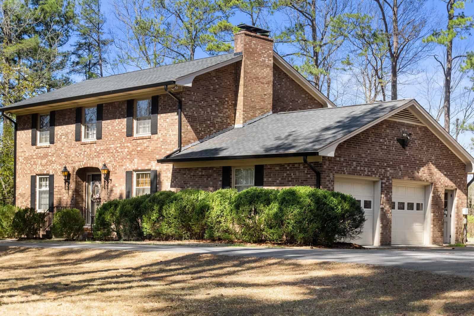 A two-story brick suburban house with a single-car garage and a landscaped front yard with bushes and bare trees under a clear sky.
