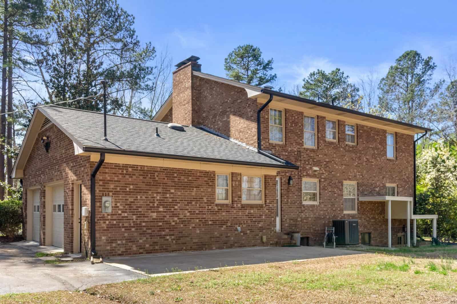 A two-story brick residential building with a black shingled roof, multiple windows, and a carport on a sunny day with trees in the background.