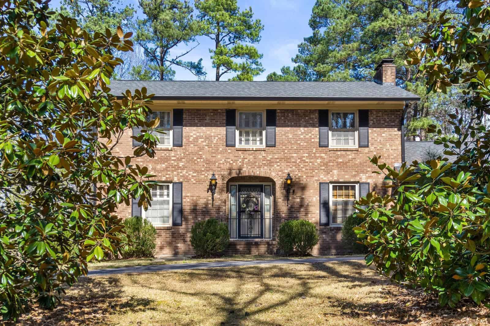 A two-story brick suburban house with a gabled roof and a landscaped front yard featuring green shrubs.