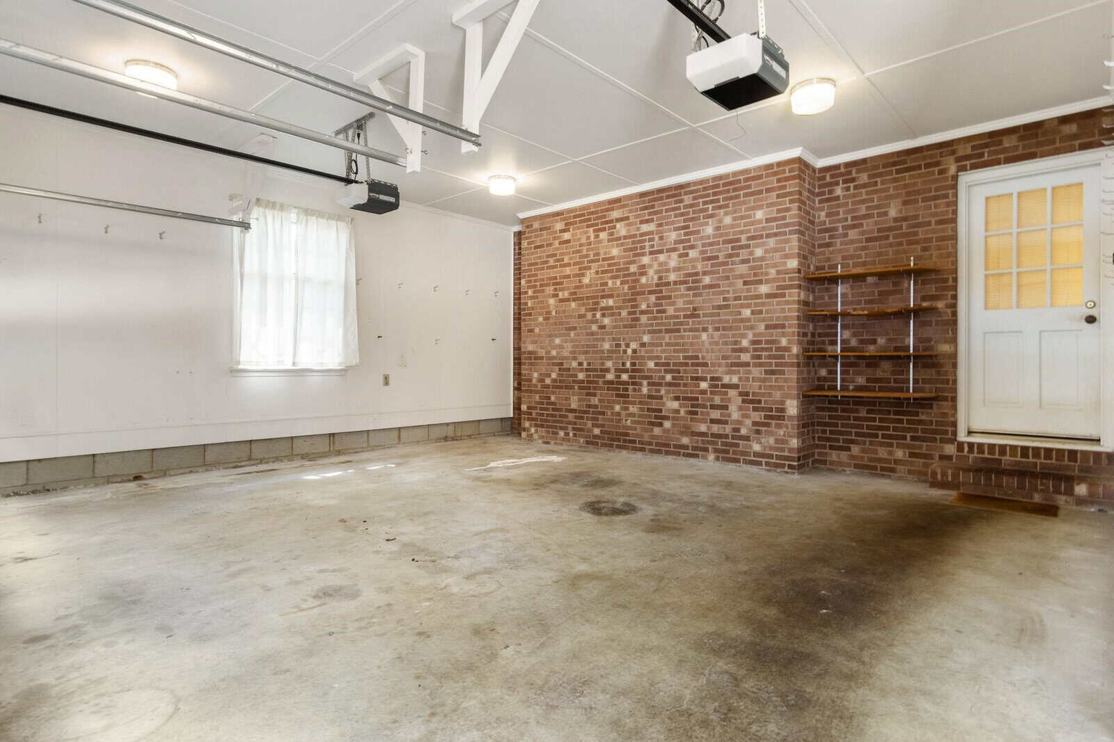 An empty garage with a concrete floor showing stains and wear, brick walls, a wooden side door, and an automatic garage door opener attached to the ceiling.