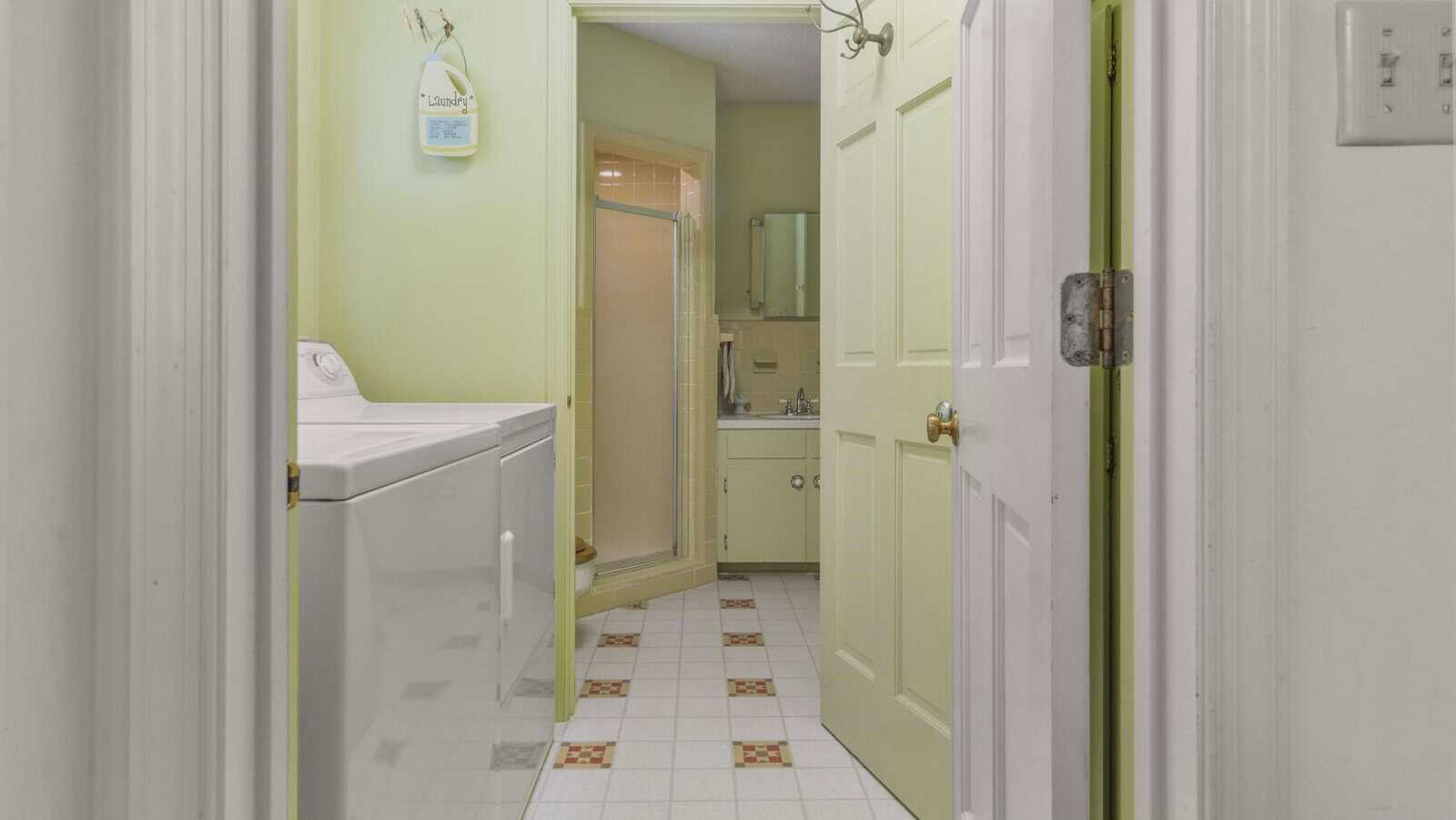 A view into a narrow laundry room with a washer on the left, leading into a bathroom with a glass shower at the end. the walls are painted light green, and the floor is tiled with a white and terracotta pattern.